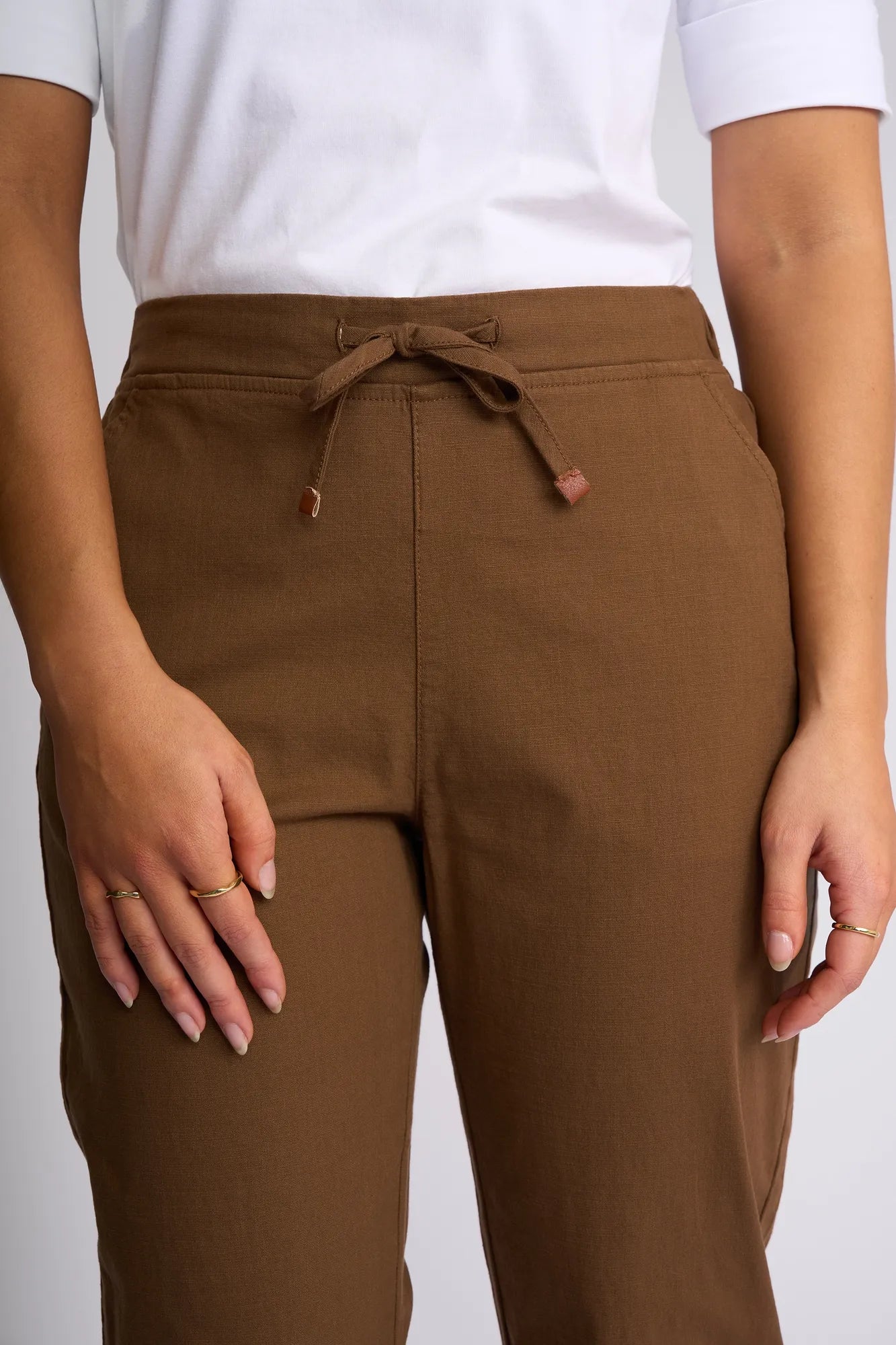 A person wearing a white shirt and Vassalli’s Denver 7/8 Pant in Pecan stands with one hand resting near a pocket. The skinny-leg pant is crafted from soft stretch cotton, and several simple rings adorn their fingers.