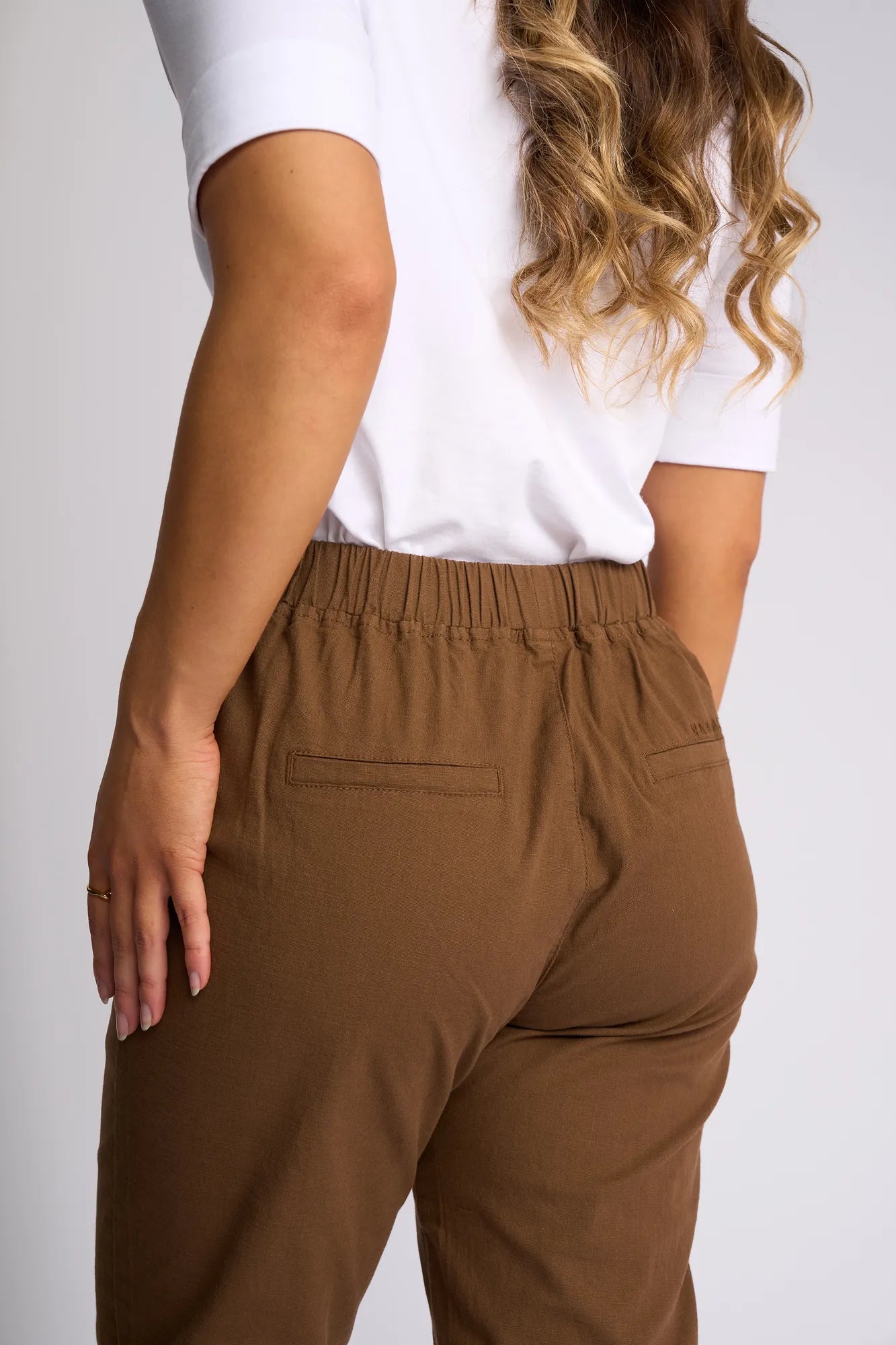 Person wearing a white short-sleeve shirt and Vassalli Denver 7/8 Pant in Pecan, shown from the back. One hand is resting on their hip. The person has wavy, light brown hair.