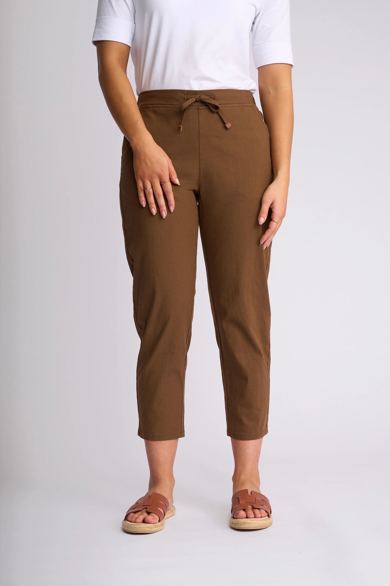 A person is seen standing against a plain light background, wearing a white short-sleeve top, the Vassalli Denver 7/8 Pant in Pecan—a stretch cotton pant featuring a skinny-leg silhouette—and brown sandals. The person’s face is not visible.