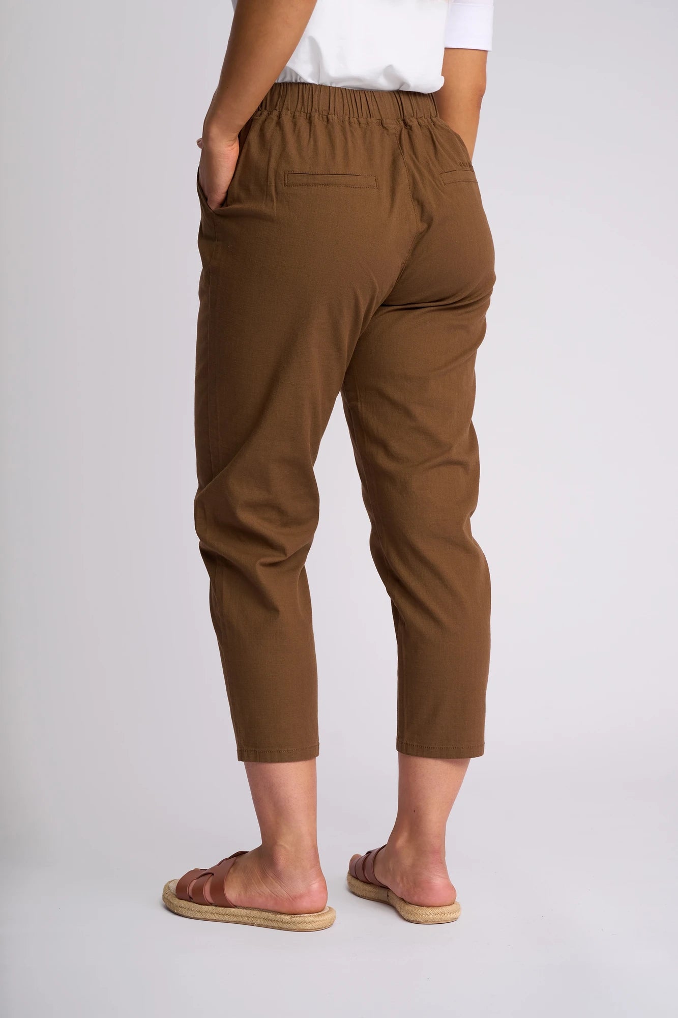 A person wearing Vassalli’s Denver 7/8 Pant in Pecan—a brown, stretch cotton skinny-leg style—paired with a white shirt and tan sandals stands with one hand in their pocket against a light background.