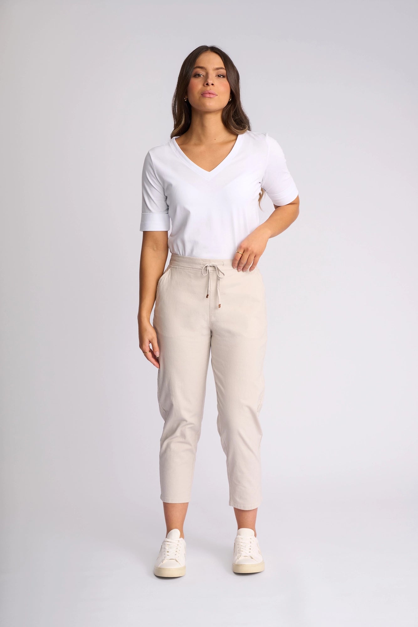 A woman stands facing forward against a plain light background, wearing a white V-neck shirt, the Vassalli Denver 7/8 Pant in Stone featuring a skinny-leg silhouette crafted from stretch cotton, and white sneakers.