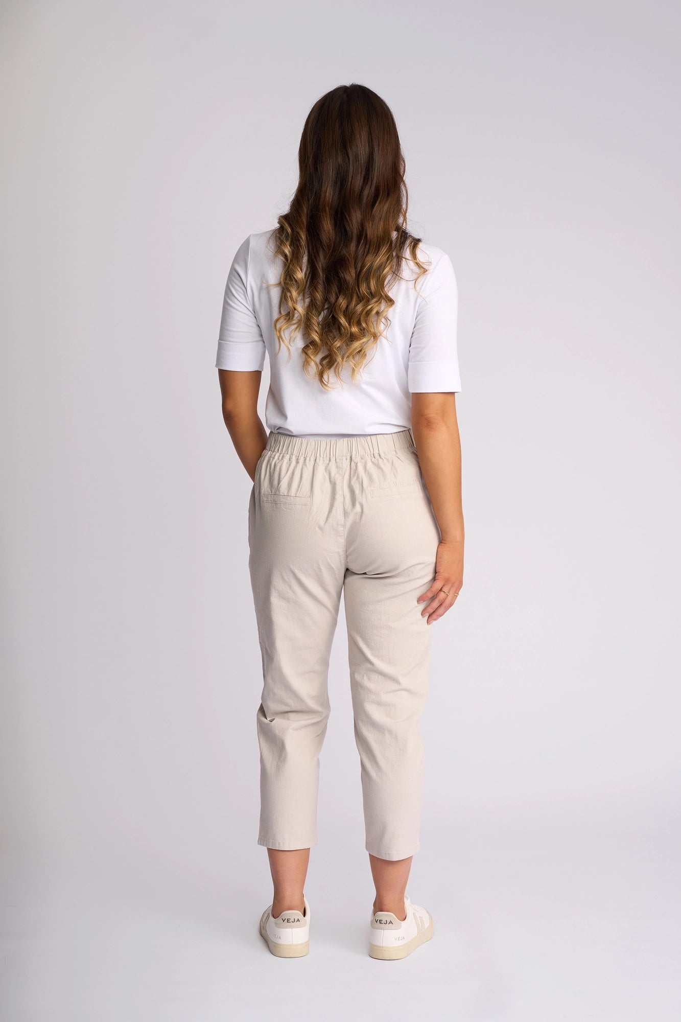 A person with long wavy brown hair stands facing away, wearing a white shirt, Vassalli Denver 7/8 Pant in Stone, and white shoes against a plain light background.