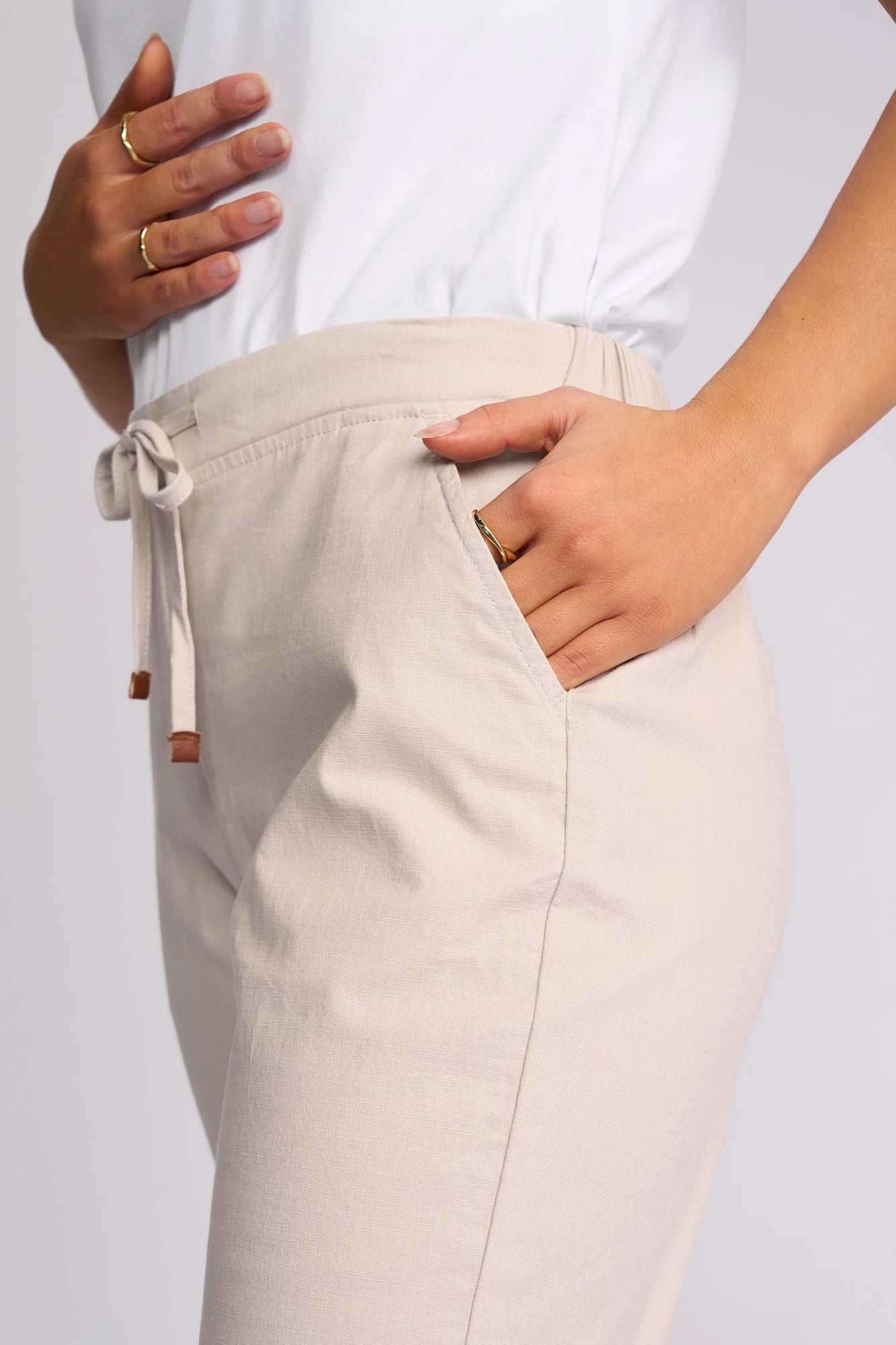 A person wearing the Vassalli Denver 7/8 Pant in Stone—a stretch cotton pant with a skinny-leg silhouette—paired with a white top, one hand in their pocket and the other resting on their waist. They are accessorized with gold rings on their fingers.