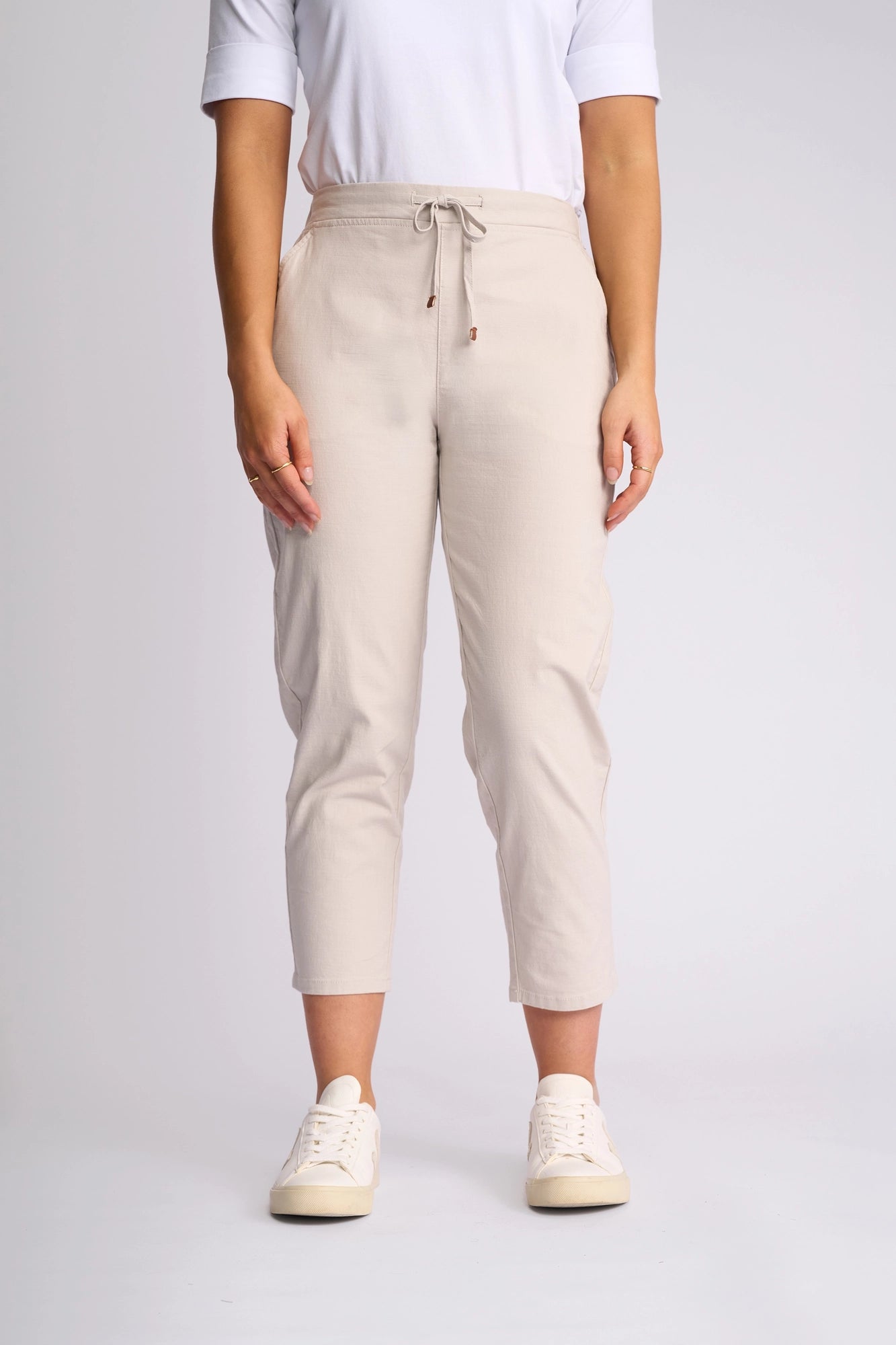 A person wearing the Vassalli Denver 7/8 Pant in Stone—an on-trend skinny-leg pant crafted from stretch cotton, featuring a drawstring waist—is paired with a white short-sleeve top and white sneakers. The individual stands against a light gray background, with their face not visible in the image.