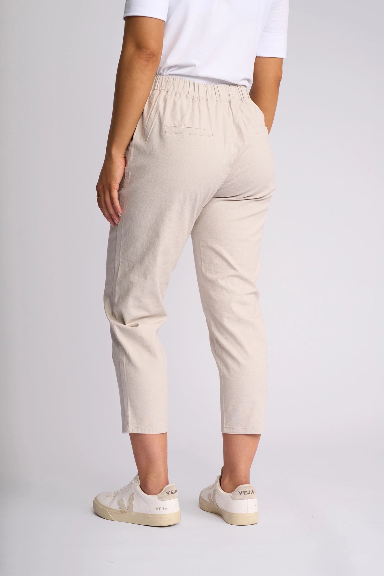A person stands facing away, wearing the Vassalli Denver 7/8 Pant in Stone—a cropped, skinny-leg pant style crafted from stretch cotton—paired with a white top and white sneakers featuring VEJA branding on the heel, against a plain light background.