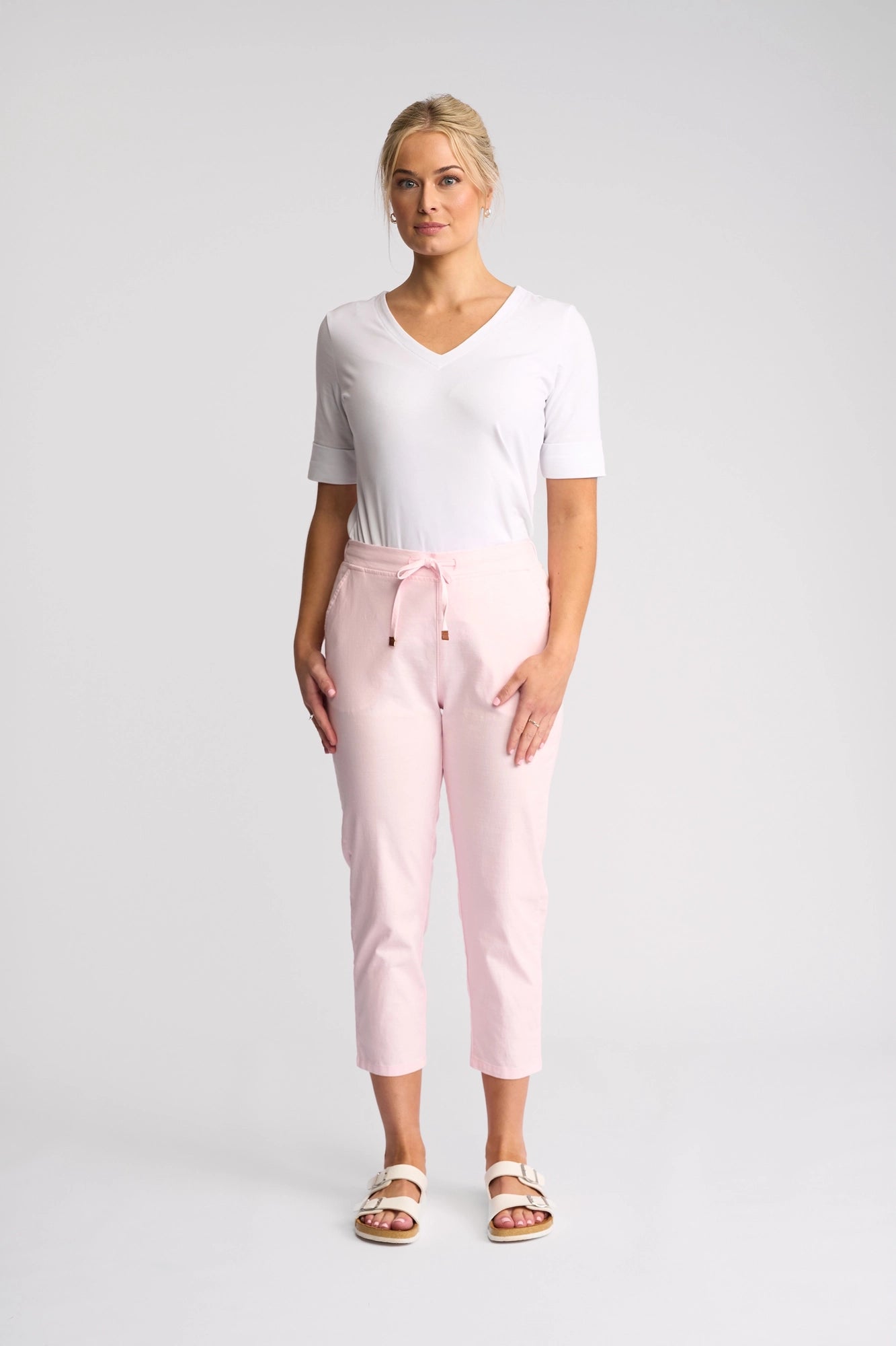 A woman stands facing forward, wearing a white V-neck top and the Vassalli Denver 7/8 Pant in Taffy, featuring a skinny-leg silhouette crafted from stretch cotton, paired with white sandals, against a plain light gray background.