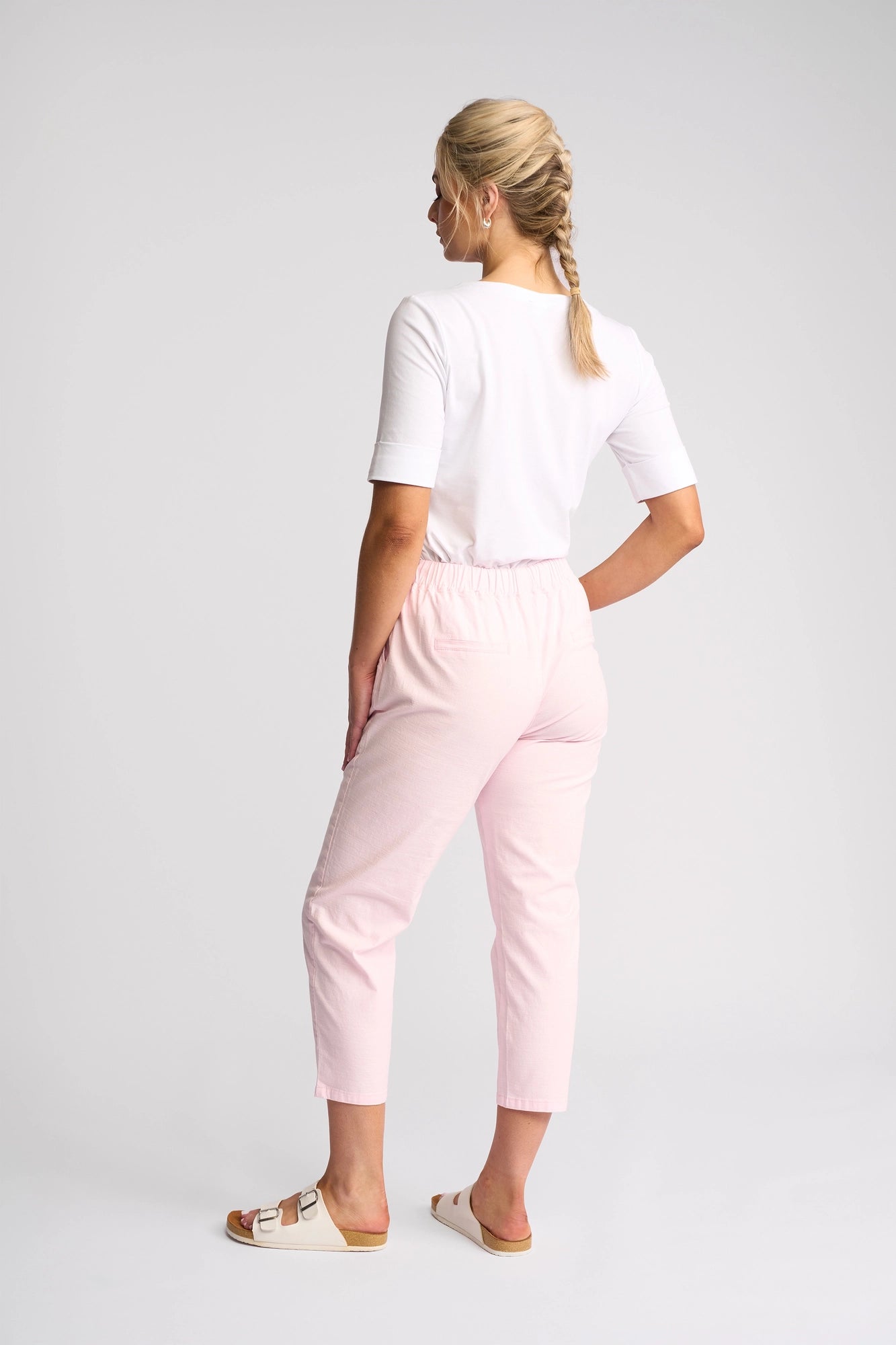 A woman with blonde hair in a braid stands with her back to the camera, wearing a white top, the Vassalli Denver 7/8 Pant in Taffy—a light pink, skinny-leg pant crafted from comfortable stretch cotton—and white sandals, all set against a plain, light background.