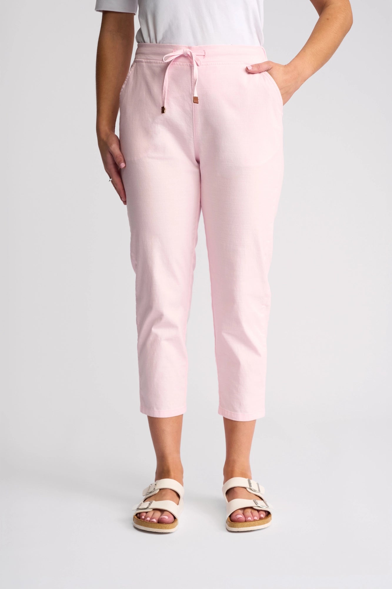 A person wearing the Vassalli Denver 7/8 Pant in Taffy—light pink, stretch cotton, skinny-leg pants—paired with a white top and white sandals stands against a plain background, one hand in their pocket and the other at their side.