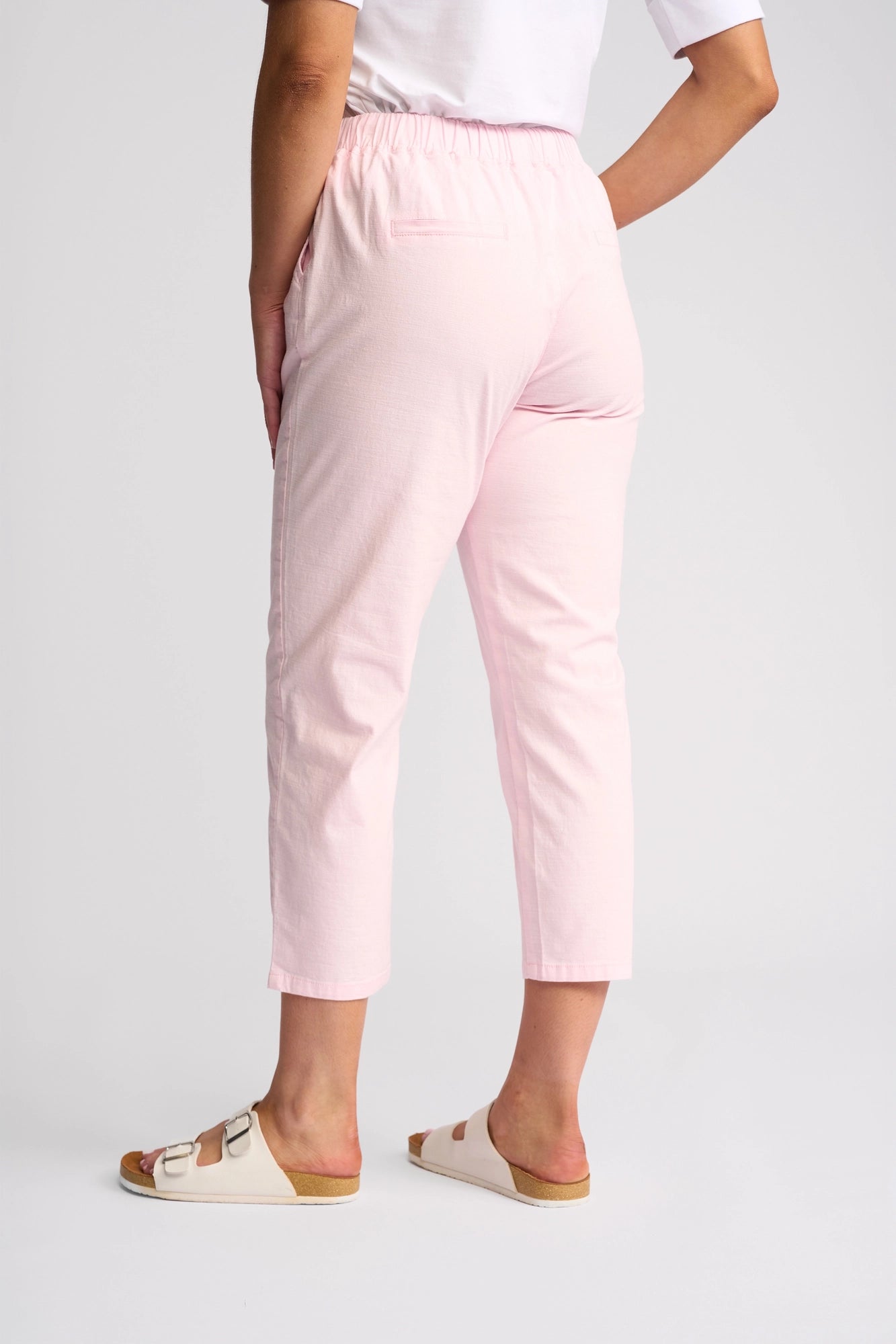 A person is shown from the back waist down, wearing Vassalli’s Denver 7/8 Pant in Taffy—a light pink, skinny-leg style made from stretch cotton—paired with a white T-shirt and white sandals, standing against a plain light background.
