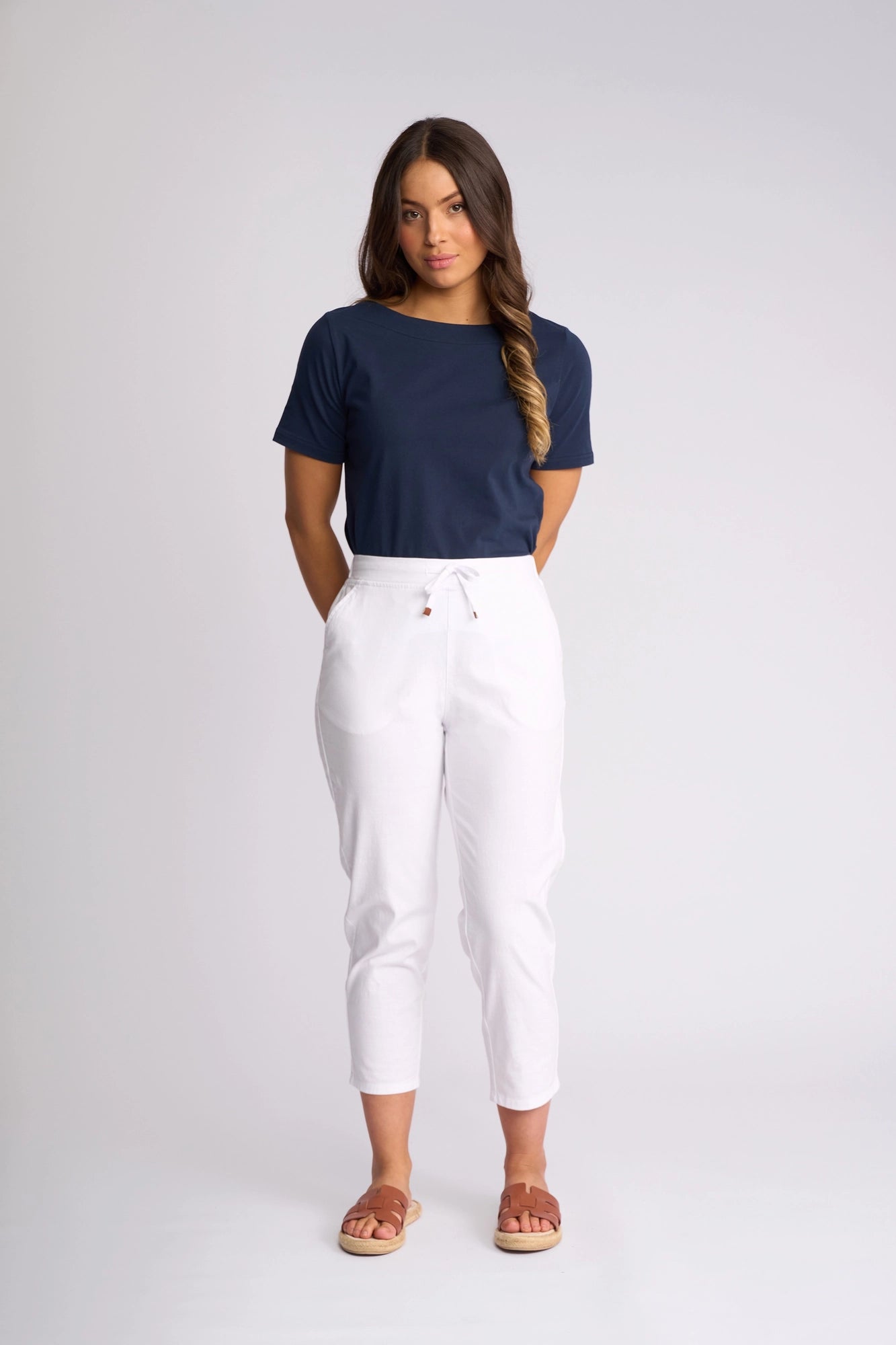 A woman stands facing forward with her hands behind her back, wearing a navy T-shirt, Vassalli Denver 7/8 Pant in white stretch cotton with a skinny-leg silhouette, and sandals. The background is plain light gray.
