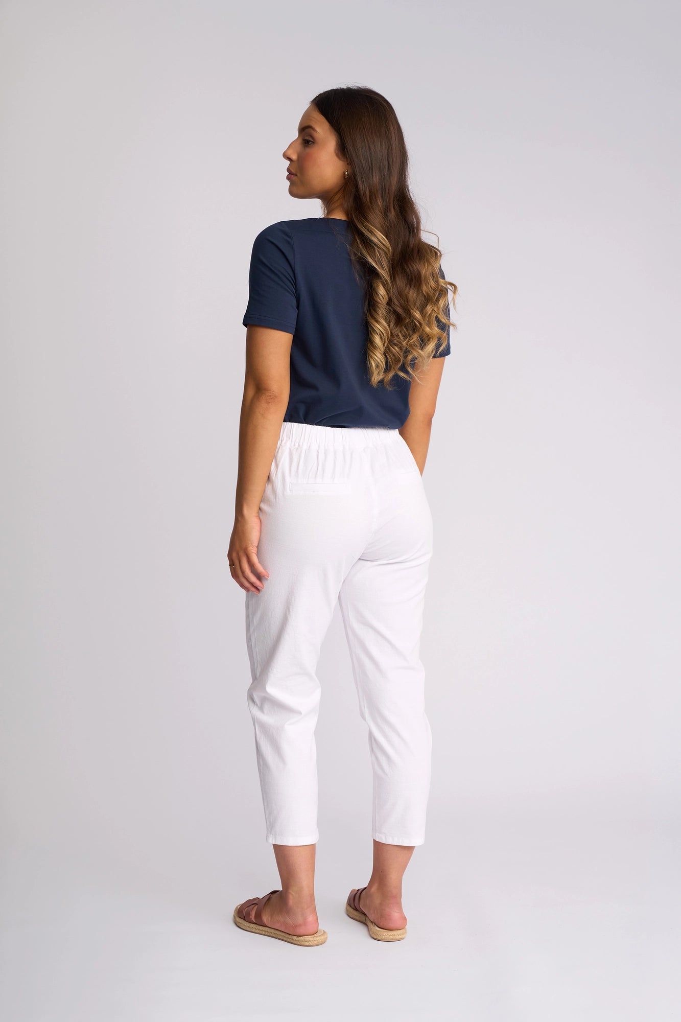 A woman with long wavy hair stands facing away, wearing a navy blue t-shirt, Vassalli Denver 7/8 Pant in white (crafted from stretch cotton), and sandals, against a plain light background.
