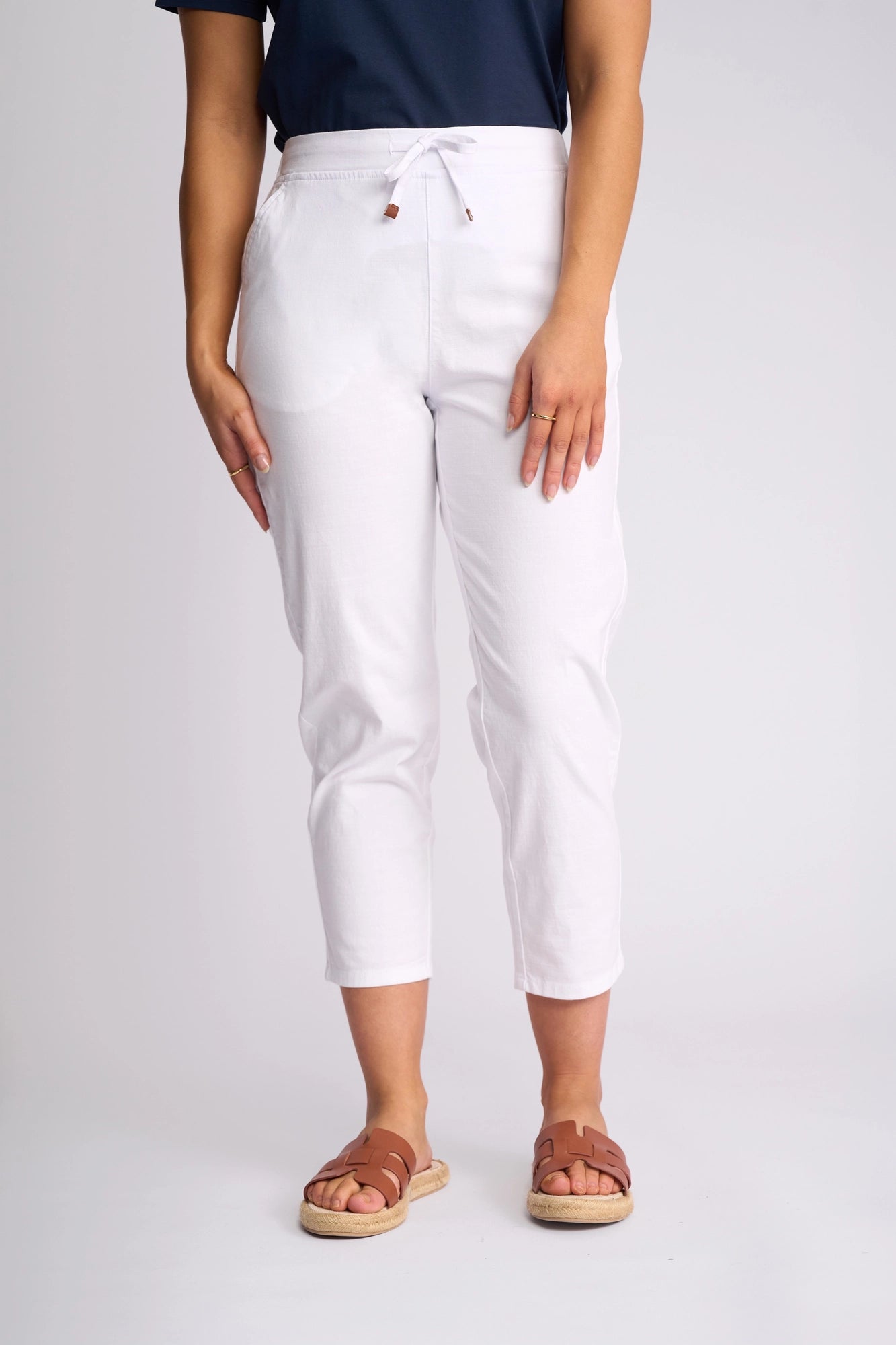 A person wearing a navy top, Vassalli Denver 7/8 Pant in white crafted from stretch cotton, and brown sandals stands against a plain light background. The image is cropped at the shoulders.