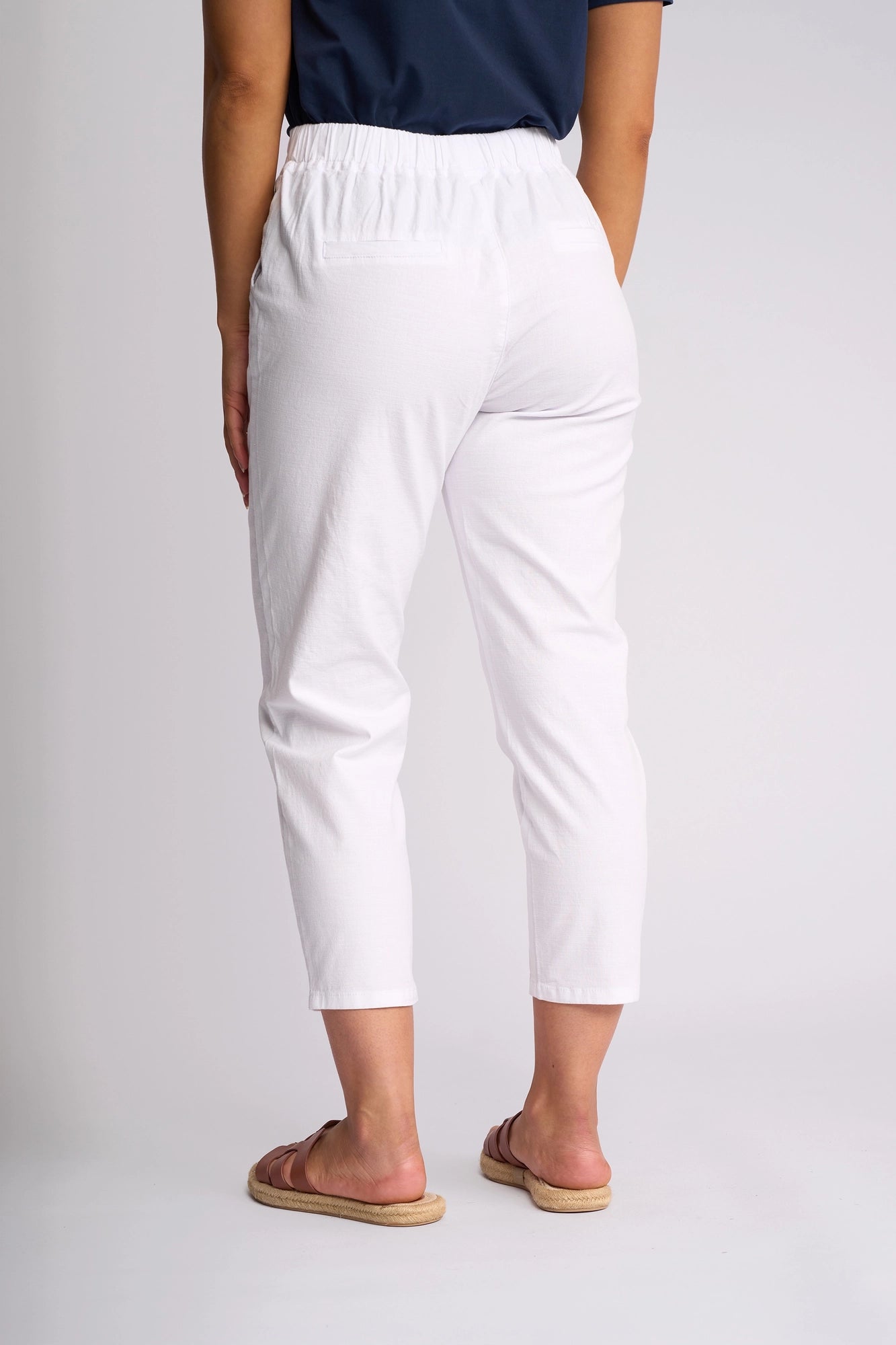 A person stands facing away, wearing a navy top and the Vassalli Denver 7/8 Pant in white, featuring a skinny-leg silhouette and an elastic waistband crafted from comfortable stretch cotton, paired with tan sandals against a plain light background.