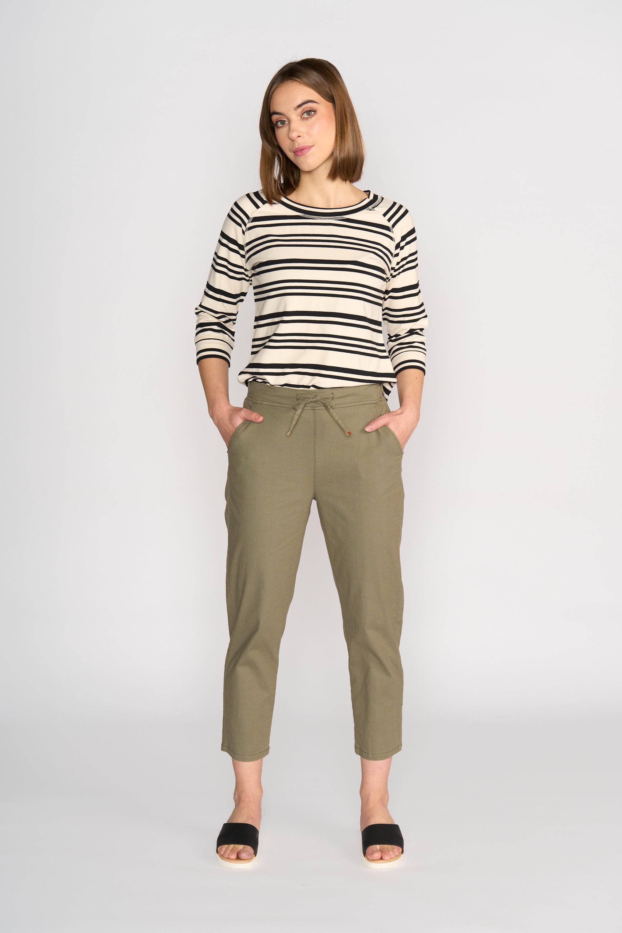A woman stands against a plain background, wearing a black and white striped long-sleeve top, Vassalli Henley 7/8 Pant - Army, and black open-toe sandals, with her hands in her pockets.