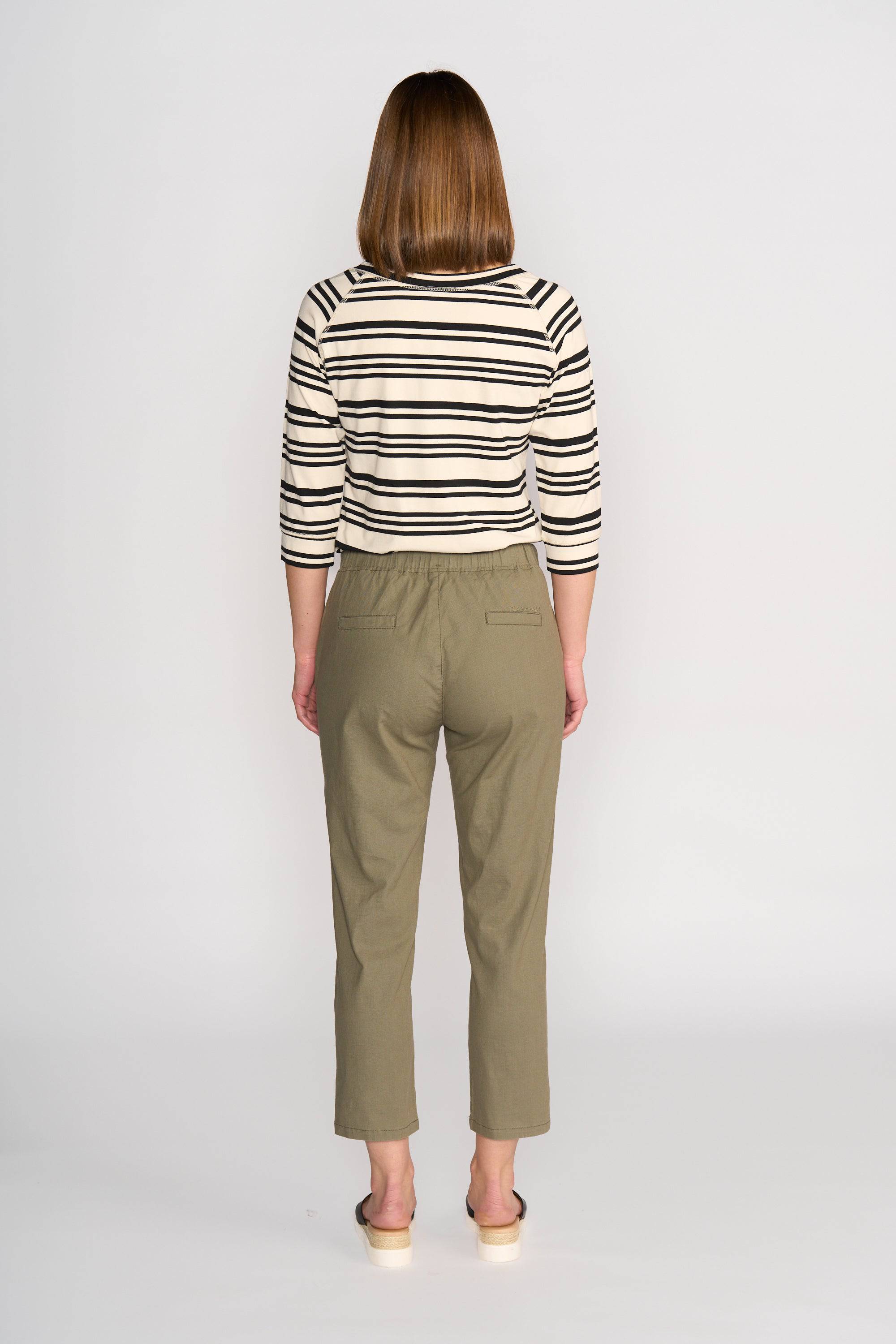 A person with straight, shoulder-length hair stands facing away, wearing a black and white striped long-sleeve shirt, Vassalli Henley 7/8 Pant - Army (olive green slim-leg stretch pants), and light slip-on shoes against a plain white background.