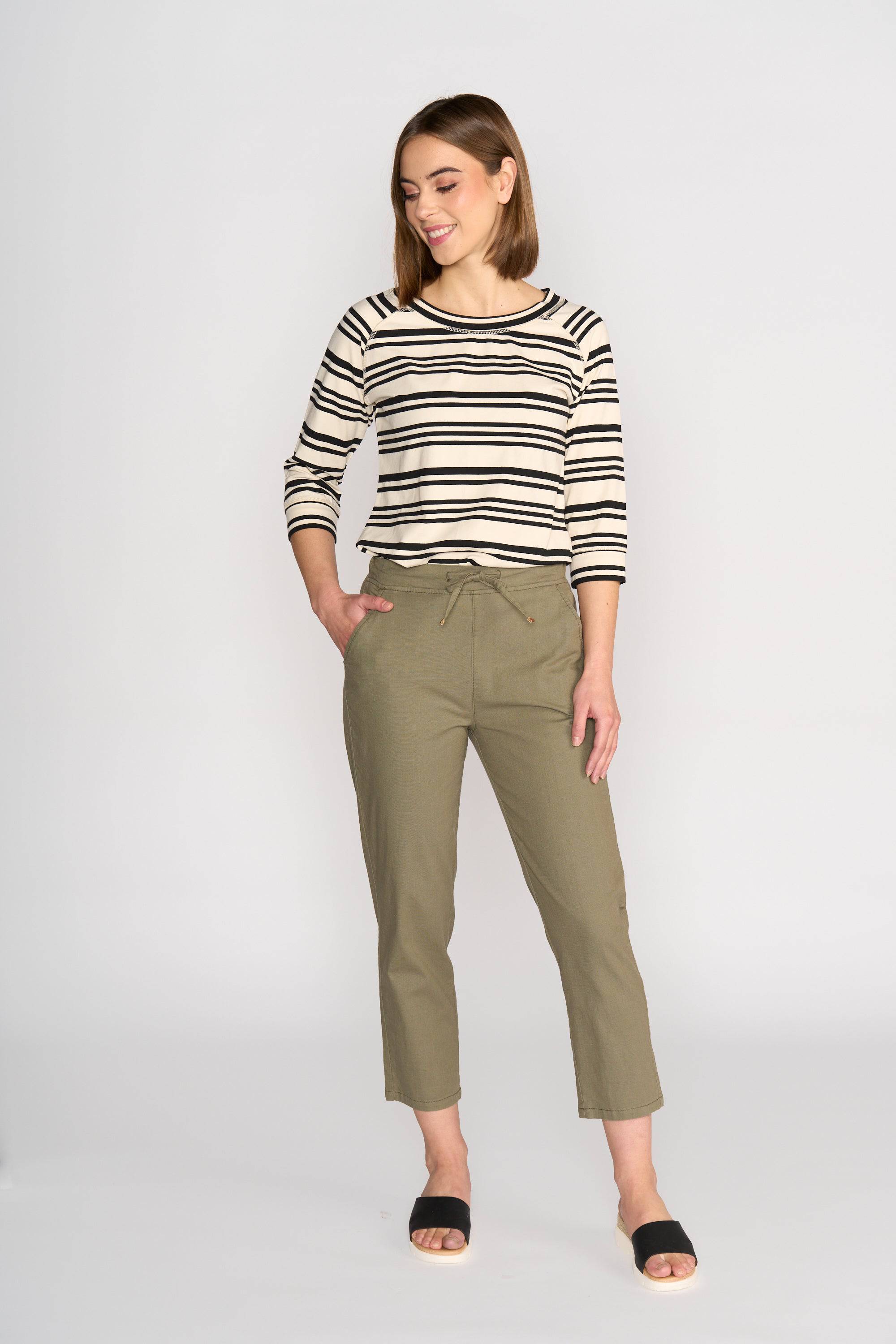 A woman smiles and looks to the side, wearing a black and white striped long-sleeve top, Vassalli's Henley 7/8 Pant in Army, and black slide sandals, against a plain light background.