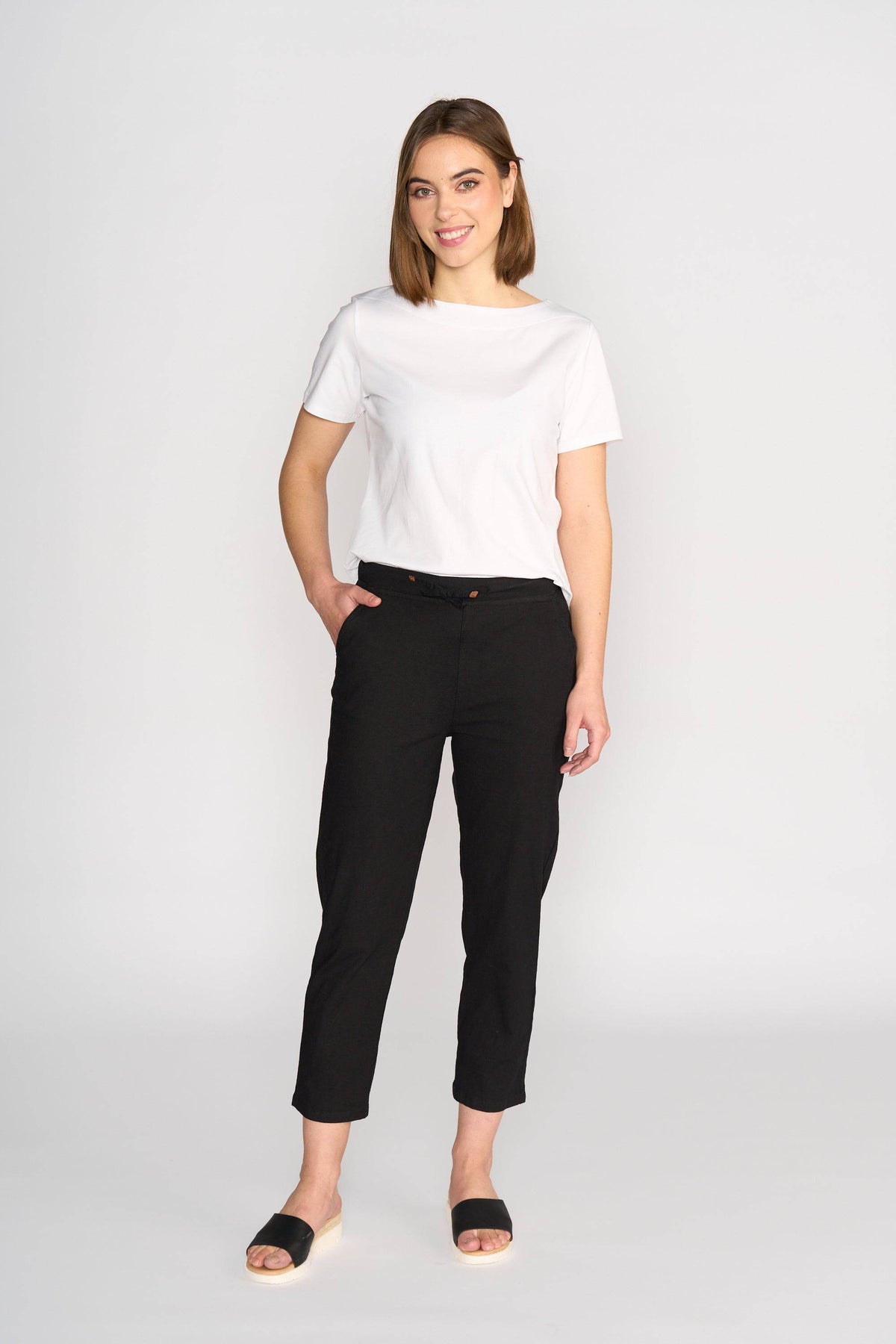 A woman stands against a white background, smiling. She wears a white short-sleeve tee and Vassalli Henley 7/8 Pant - Black, featuring an elastic waistband and slim leg, paired with black slip-on sandals. One hand is in her pocket.