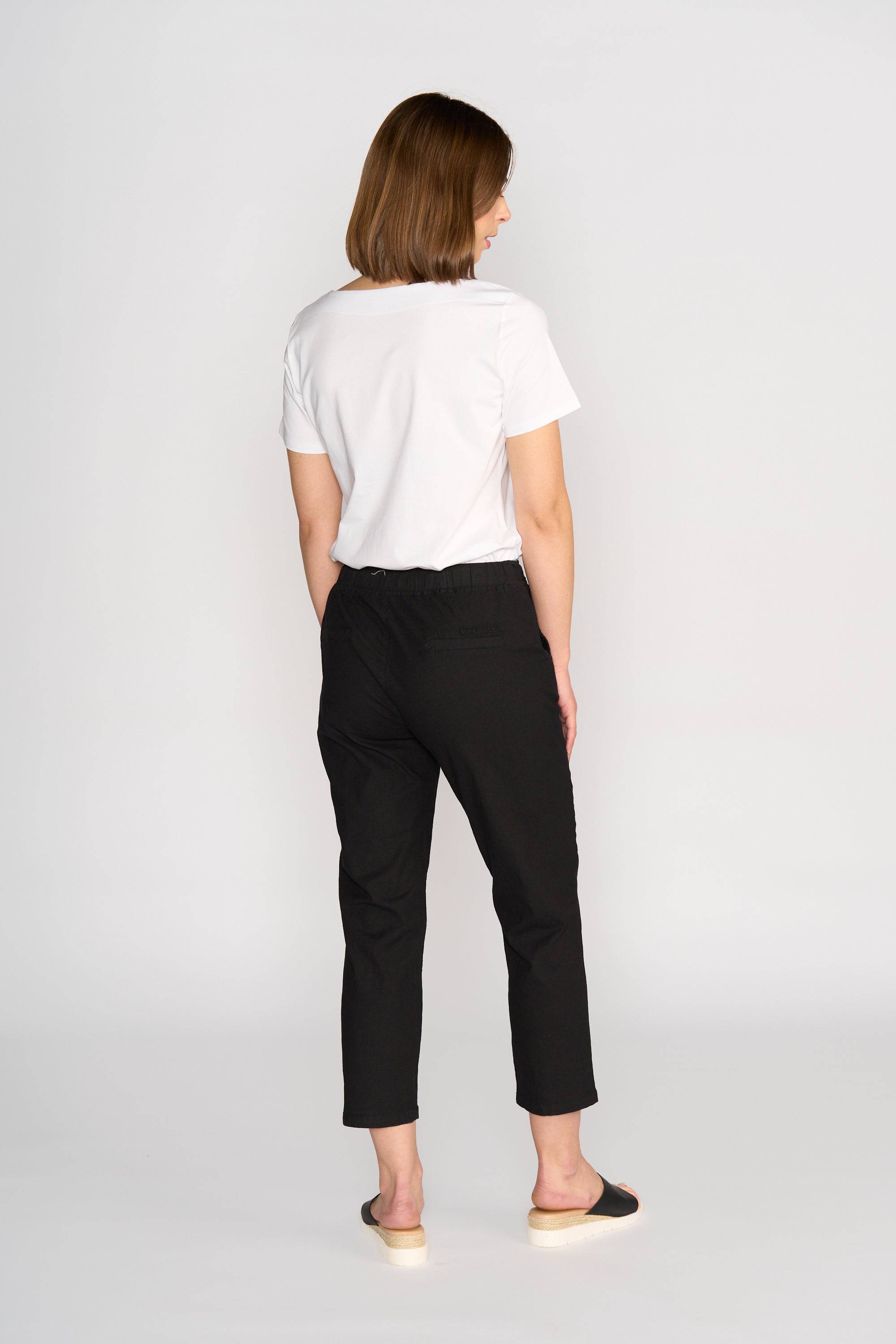 A woman with short brown hair faces away, wearing a white t-shirt and Vassalli Henley 7/8 Pant in black with an elastic waistband and black slip-on shoes, set against a plain white background.