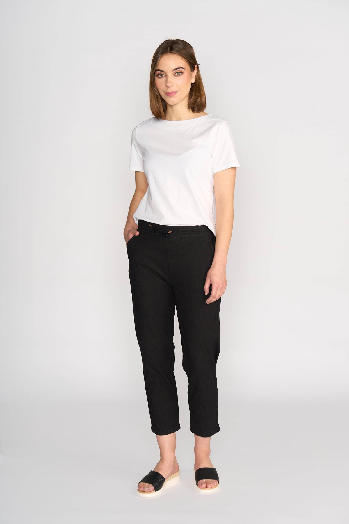 A woman with shoulder-length brown hair stands against a plain light background, wearing the Vassalli Henley 7/8 Pant in black and a white t-shirt. One hand is in her pocket as she looks at the camera with a slight smile.