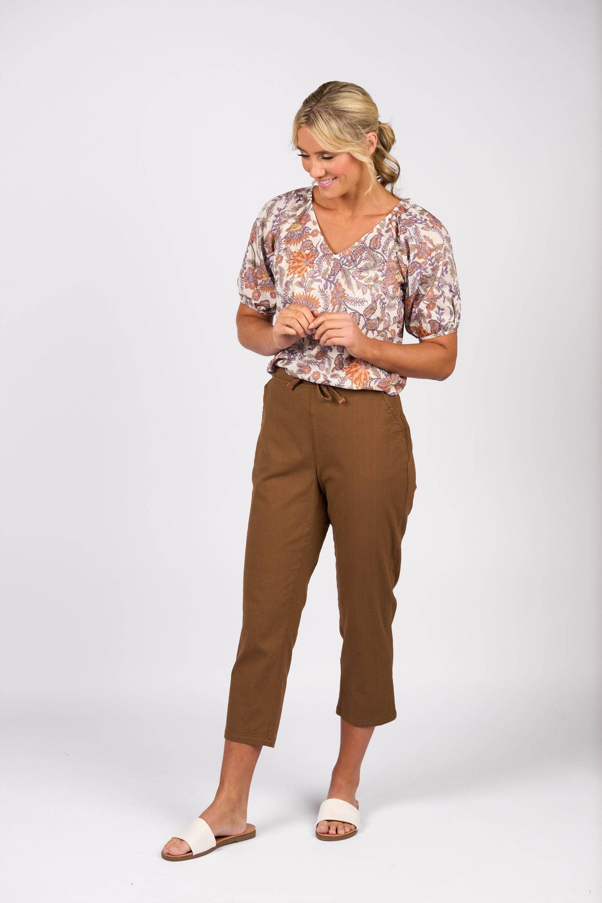A woman stands against a white background, smiling and looking down. She wears a patterned blouse, white slide sandals, and Vassalli's Henley 7/8 Pant in Pecan.