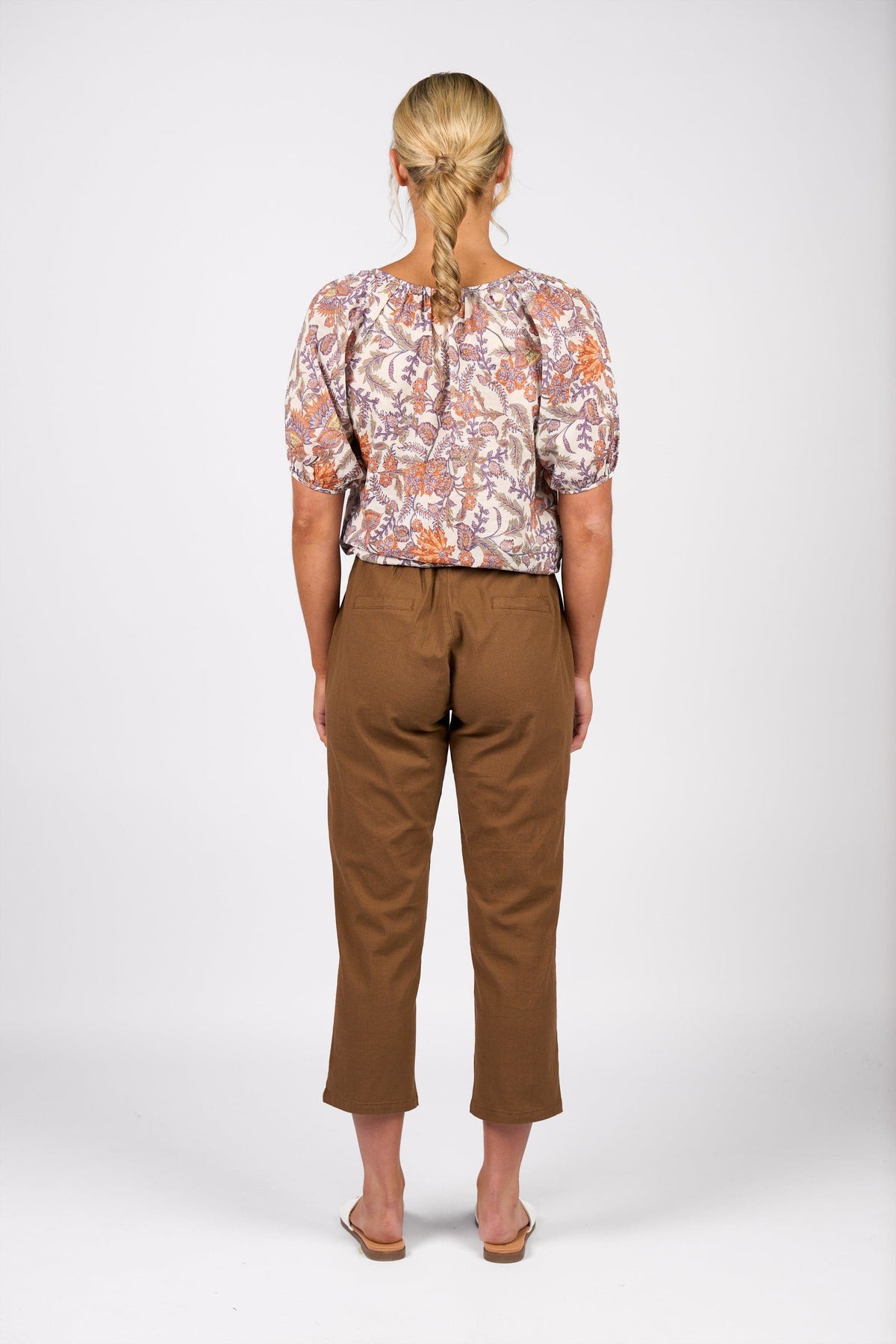 A person with blonde braided hair, shown from the back, wears a floral short-sleeve blouse, Vassalli Henley 7/8 Pant in Pecan, and sandals against a plain white background.
