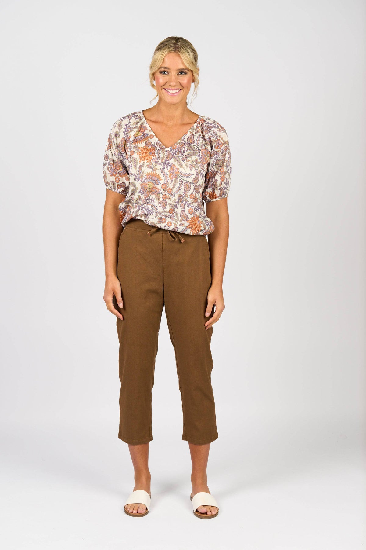 A woman smiles against a white background, wearing a floral short-sleeve blouse, Vassalli Henley 7/8 Pant in Pecan with an elastic waistband, and white sandals.