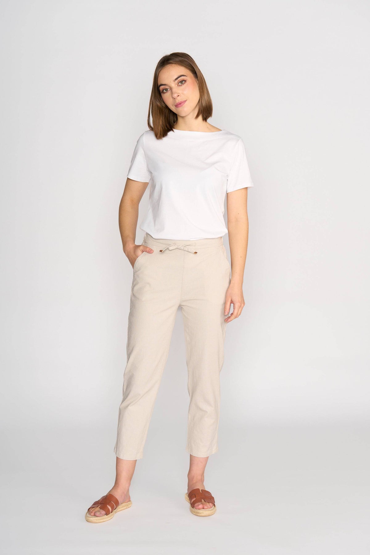 A woman stands against a plain white background wearing Vassalli’s Henley 7/8 Pant in Stone, a pair of beige slim-leg pants with an elastic waistband, paired with a white t-shirt and brown sandals. She poses with one hand in her pocket.