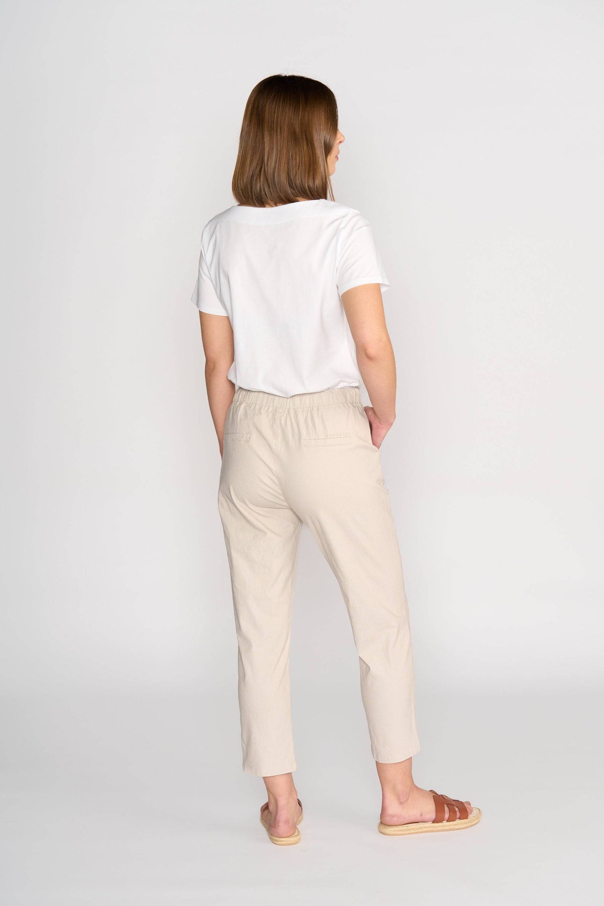 A woman faces away from the camera, wearing a white T-shirt and Vassalli Henley 7/8 Pant in Stone with an elastic waistband, paired with brown sandals, set against a plain light background.