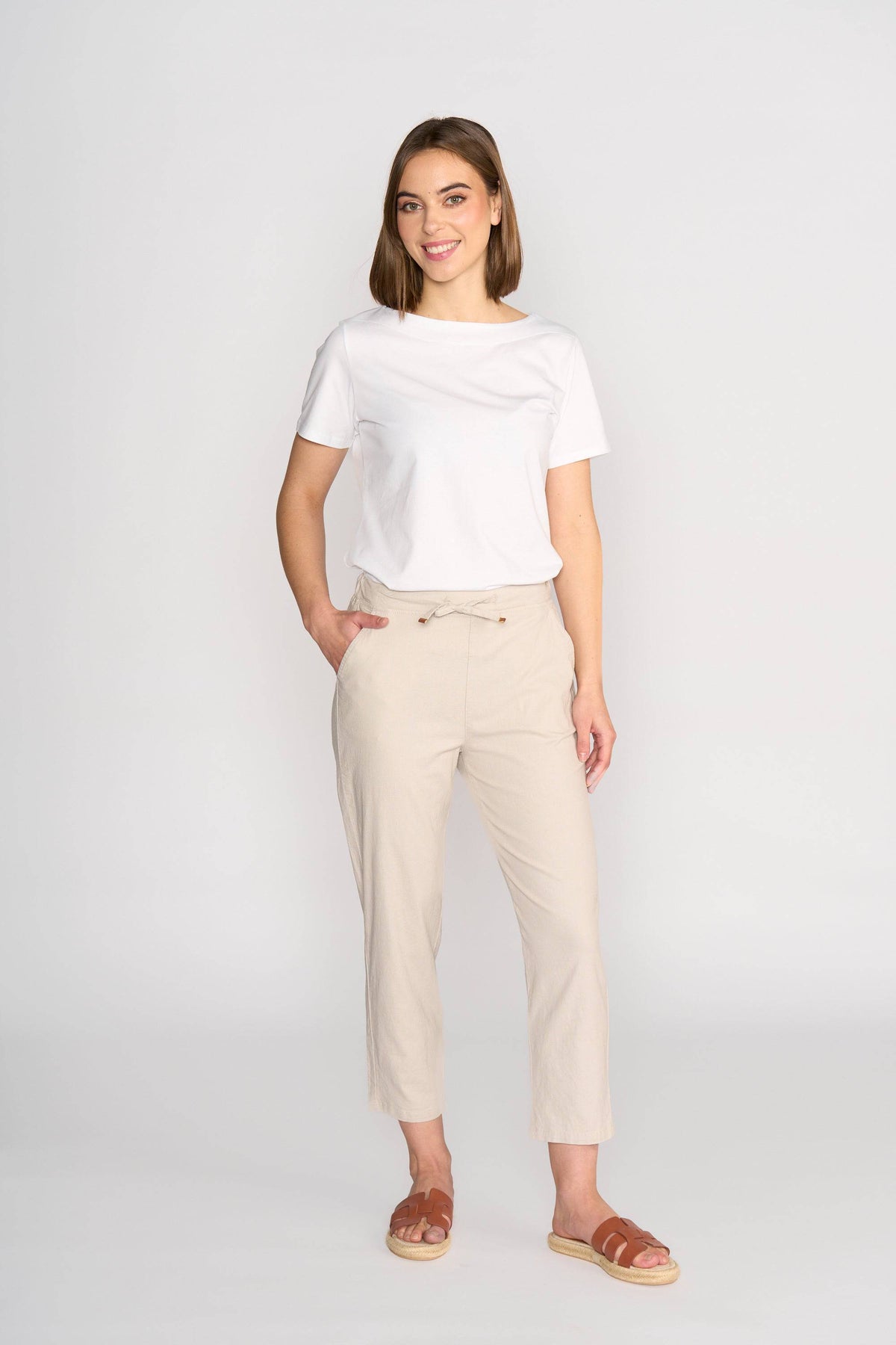 A woman with chin-length hair smiles against a light background, wearing a white t-shirt, brown sandals, and Vassalli's Henley 7/8 Pant in Stone—a slim-leg, light beige style with an elastic waistband and drawstring. Her left hand is in her pocket.