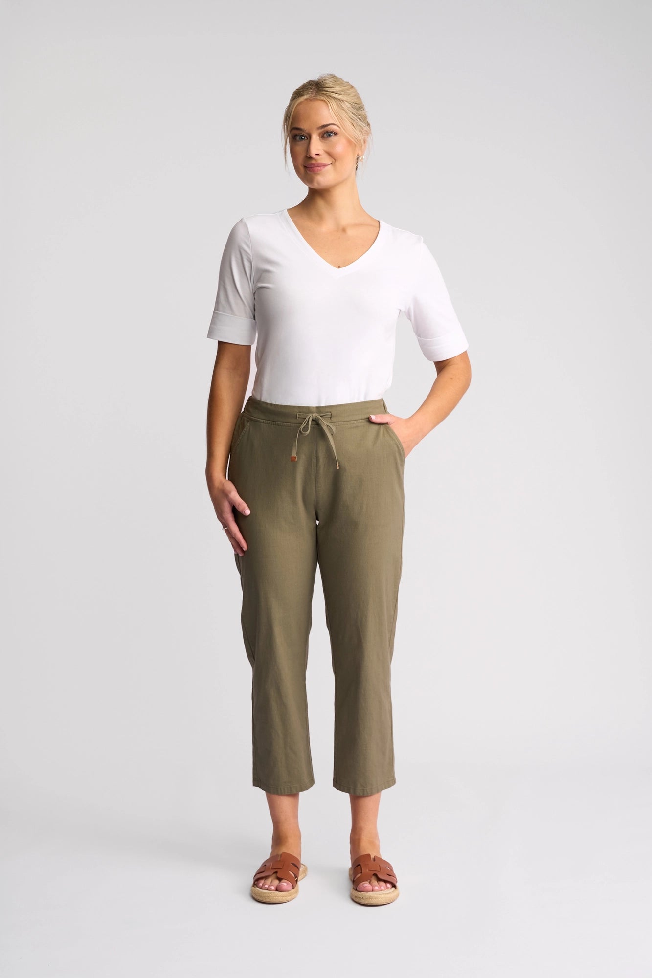 A woman stands facing forward against a plain light background, wearing a white V-neck short sleeve top, the Vassalli Henley 7/8 Pant in Army with a stretch cotton slim-leg silhouette, and brown sandals, with one hand in her pocket.