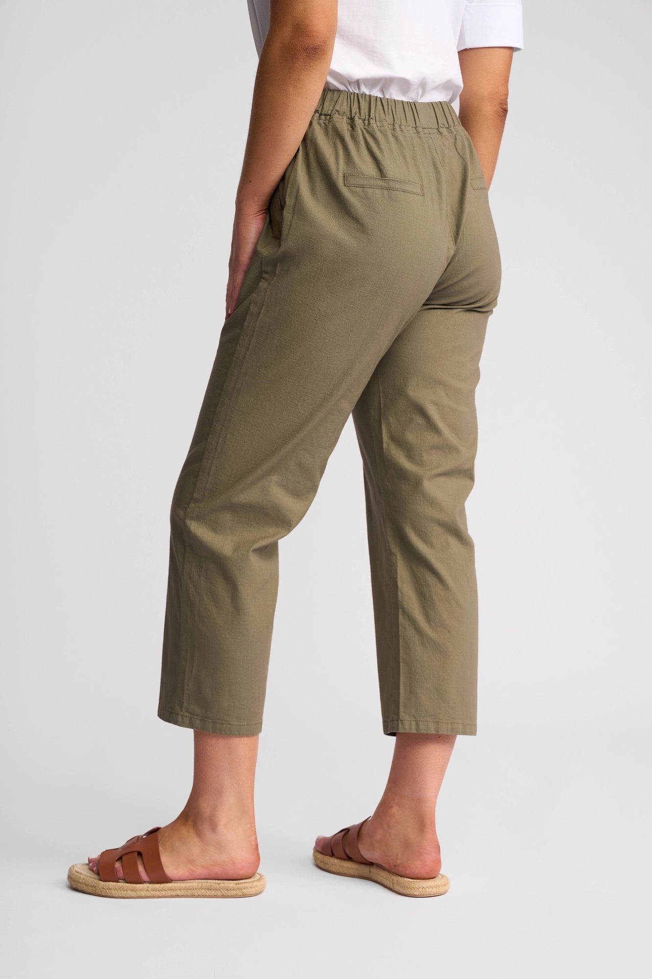 A person is shown from the back, standing against a plain white background, wearing Vassalli’s Henley 7/8 Pant in Army with a white shirt and brown sandals.