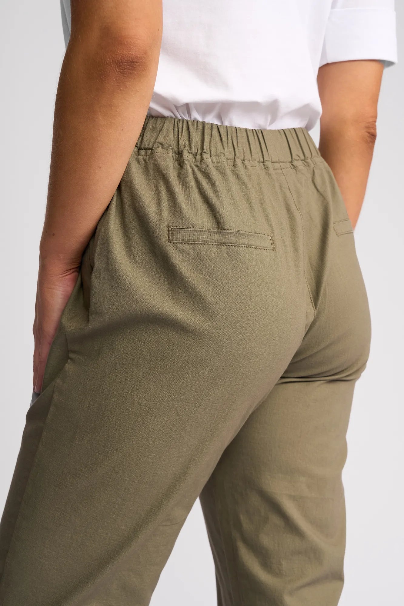 A person wearing the Vassalli Henley 7/8 Pant - Army, featuring an elastic waistband, is seen from the back with one hand in the pocket and a white shirt. The slim-leg pant is crafted from stretch cotton for all-day comfort and effortless style.