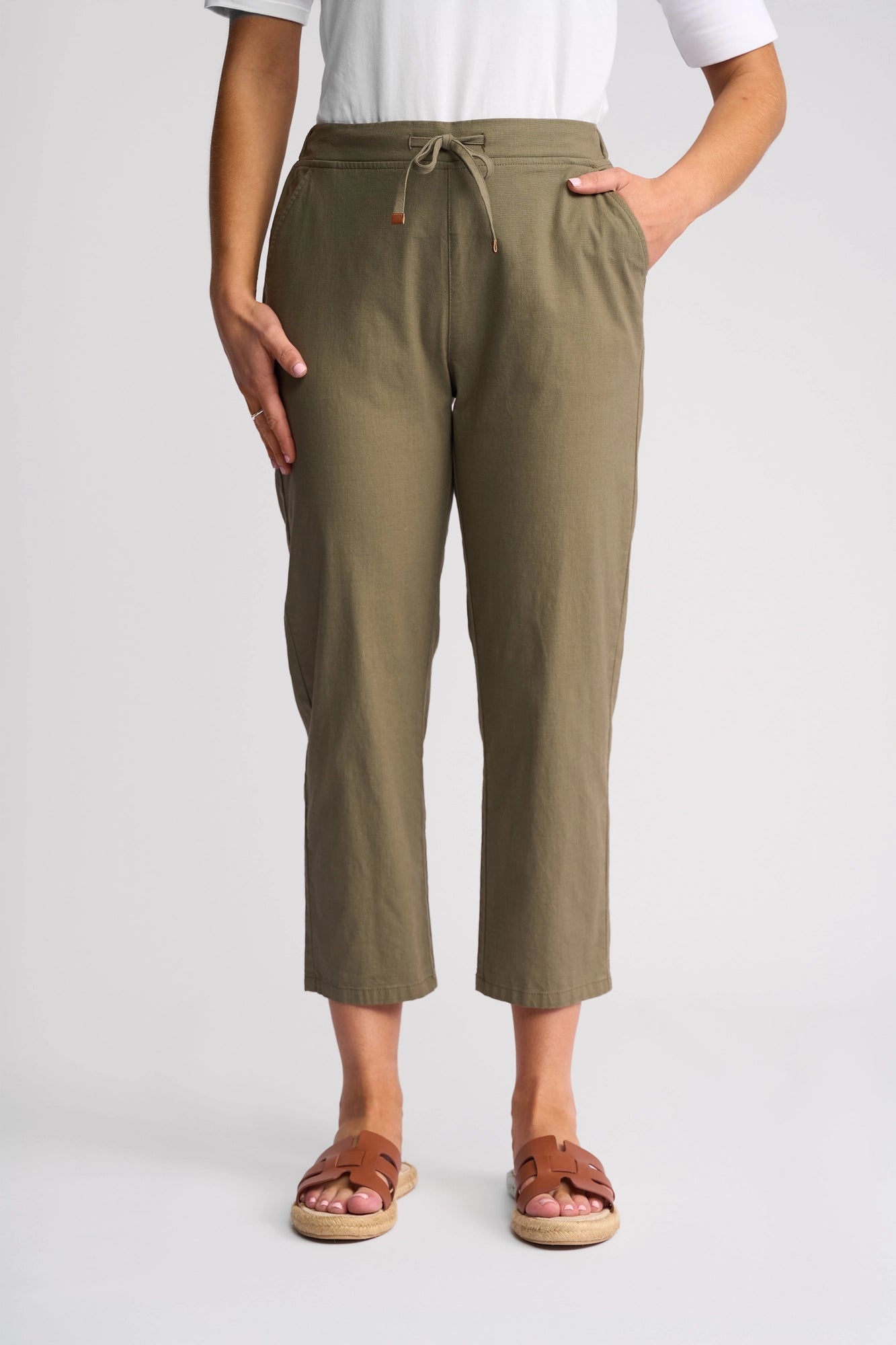 A person wearing the Vassalli Henley 7/8 Pant in Army—a slim-leg silhouette crafted from comfortable stretch cotton—paired with a white shirt and brown sandals, stands with one hand in their pocket against a plain light background.