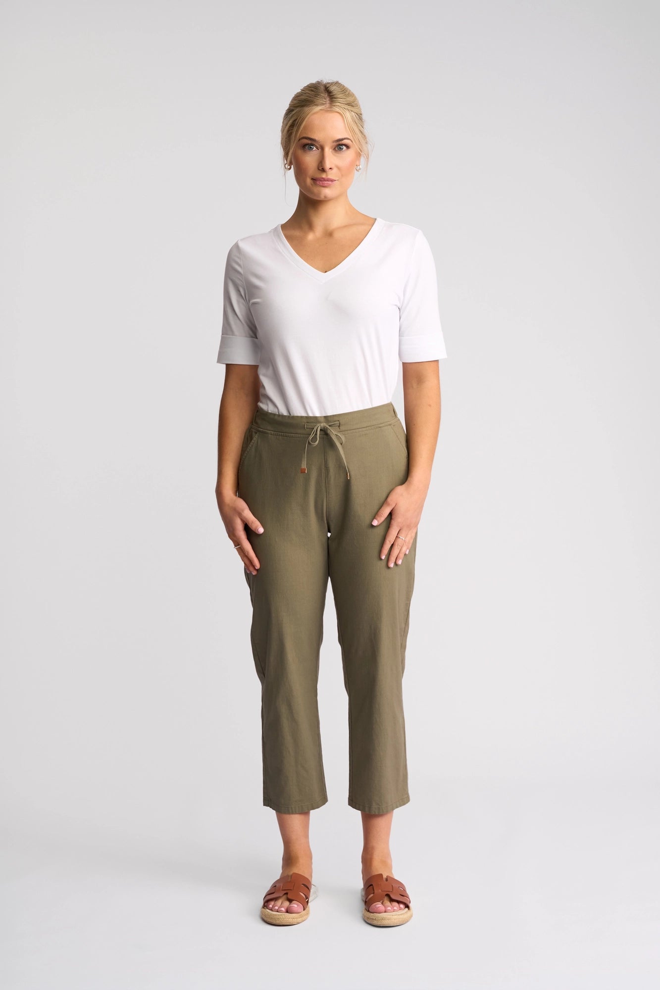 A person stands facing forward against a plain background, wearing a white short-sleeve V-neck shirt, the Vassalli Henley 7/8 Pant - Army in stretch cotton with a drawstring waist, and brown sandals.
