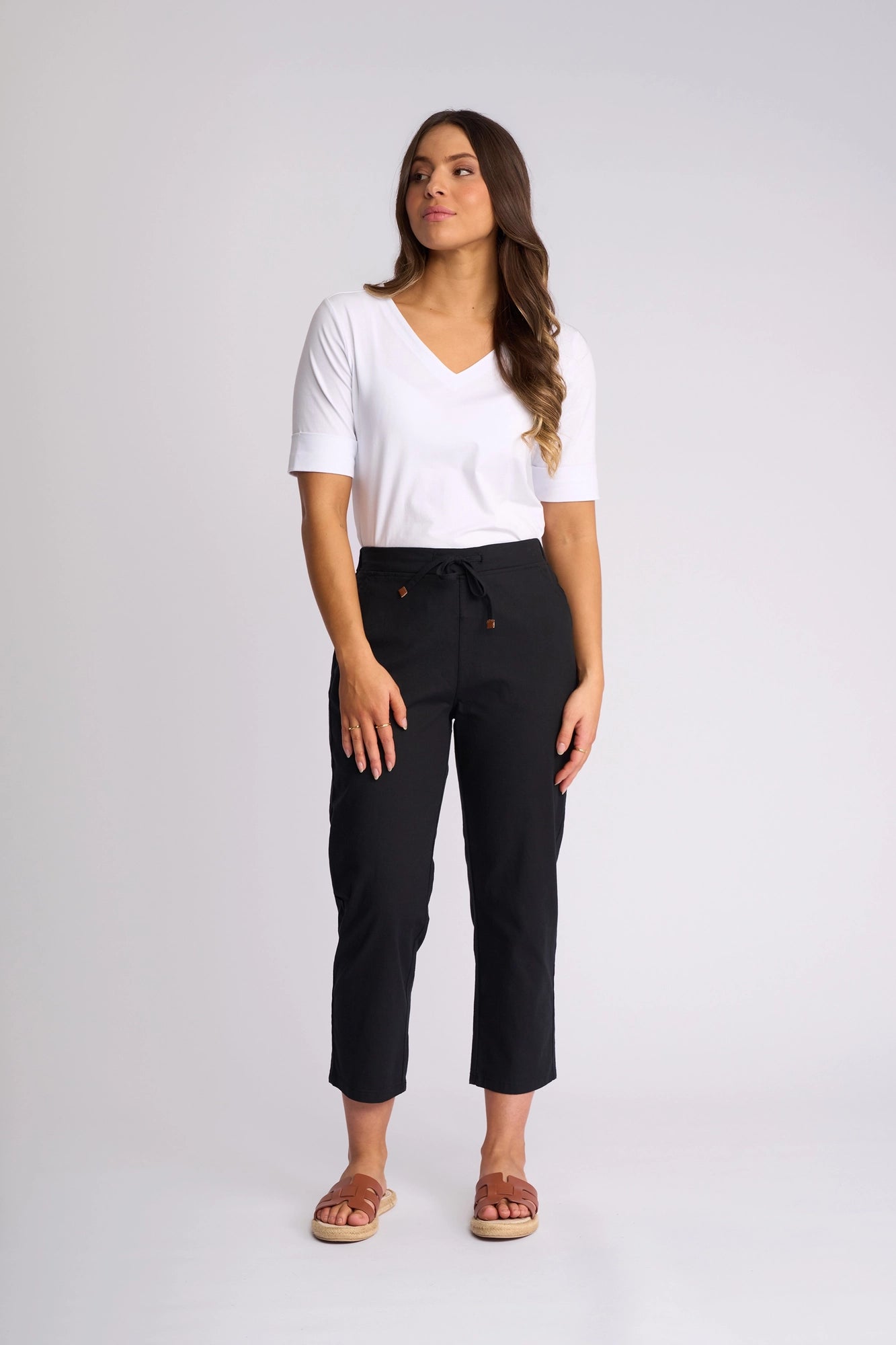 A woman stands against a plain light background, wearing a white V-neck top and the Vassalli Henley 7/8 Pant in black—crafted from stretch cotton and featuring a drawstring waist—paired with brown slip-on sandals. She gazes slightly to her left with one hand resting at her side.