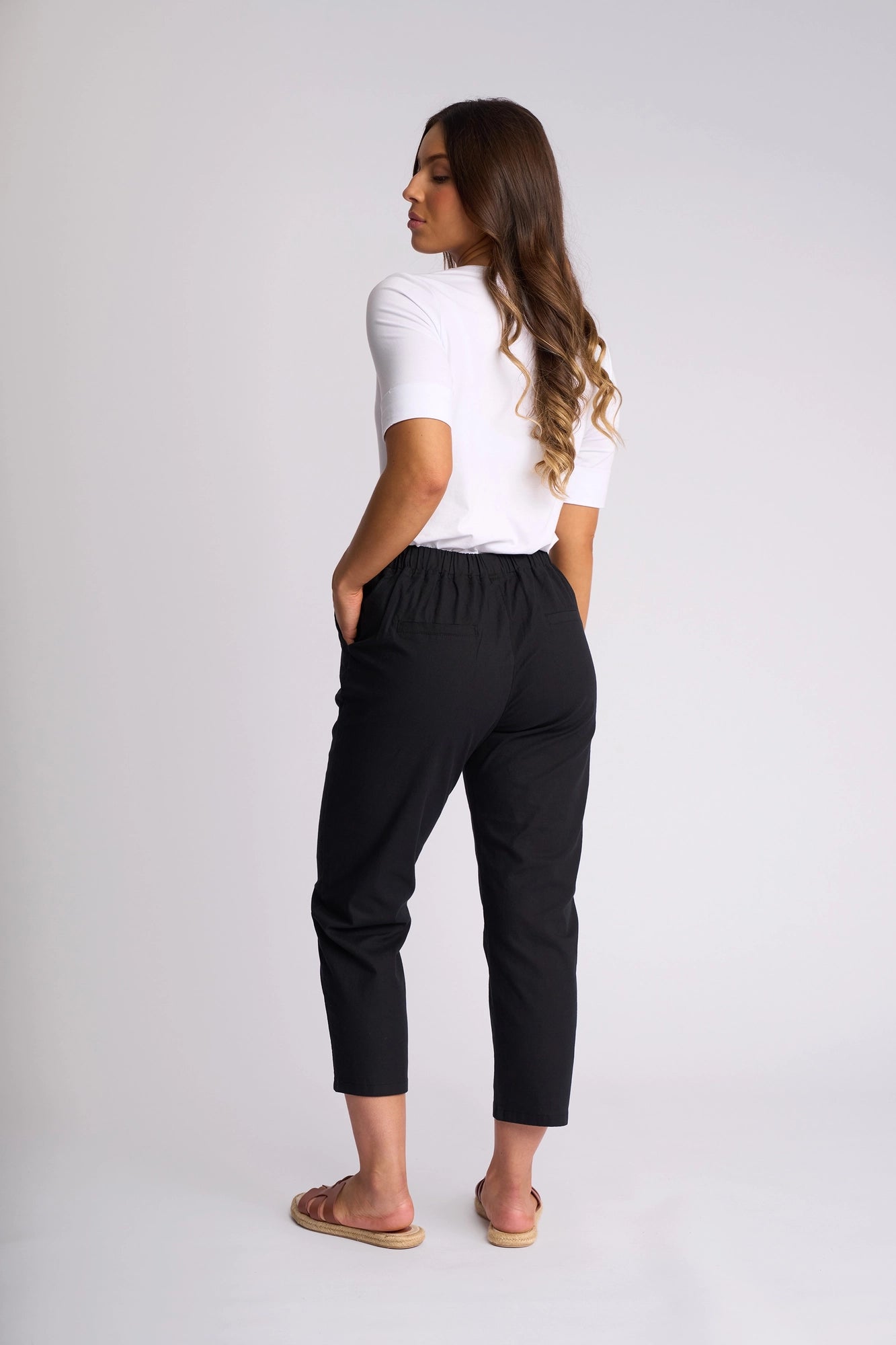 A woman with long, wavy brown hair stands with her back turned against a plain background, hands in her pockets. She is wearing a white short-sleeve top, sandals, and the Vassalli Henley 7/8 Pant in black—slim-leg pants crafted from stretch cotton.