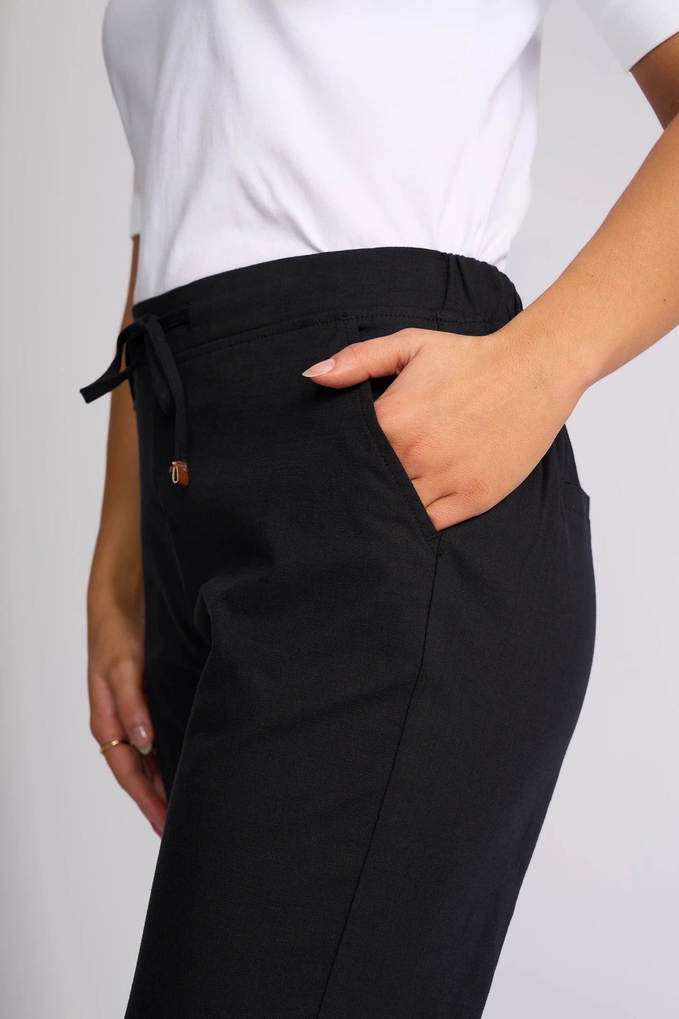 A person wearing a white shirt and Vassalli’s Henley 7/8 Pant in black stands with one hand in their pocket, showcasing a casual look from the waist to mid-thigh.