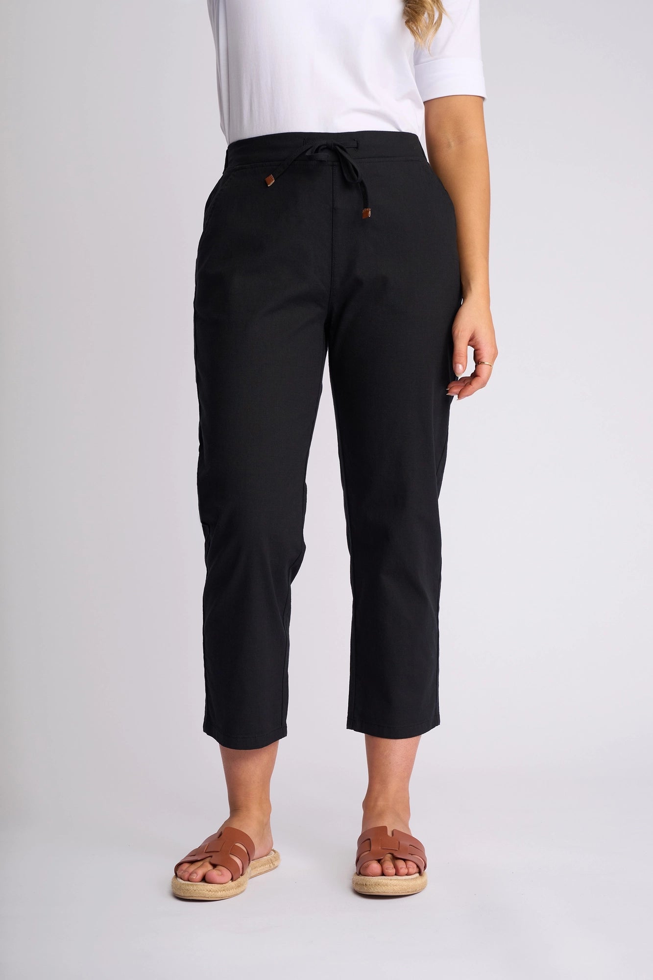 A person wears the Vassalli Henley 7/8 Pant in black, made from stretch cotton, paired with a white short-sleeve top and brown sandals, standing against a plain light background.