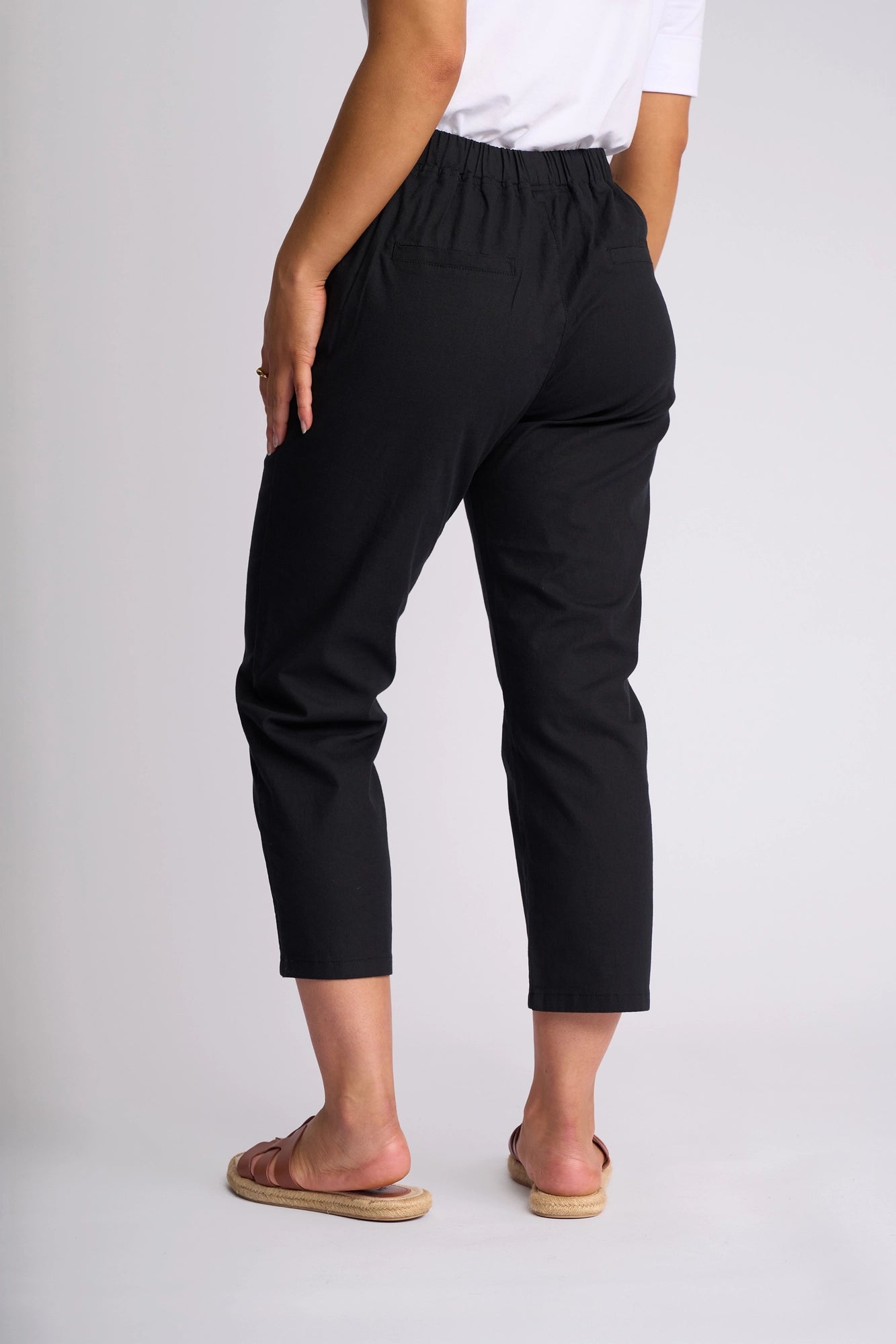 A person stands facing away against a plain light background, showcasing the back view of the Vassalli Henley 7/8 Pant in Black—crafted from stretch cotton—paired with a white top and tan flat sandals.