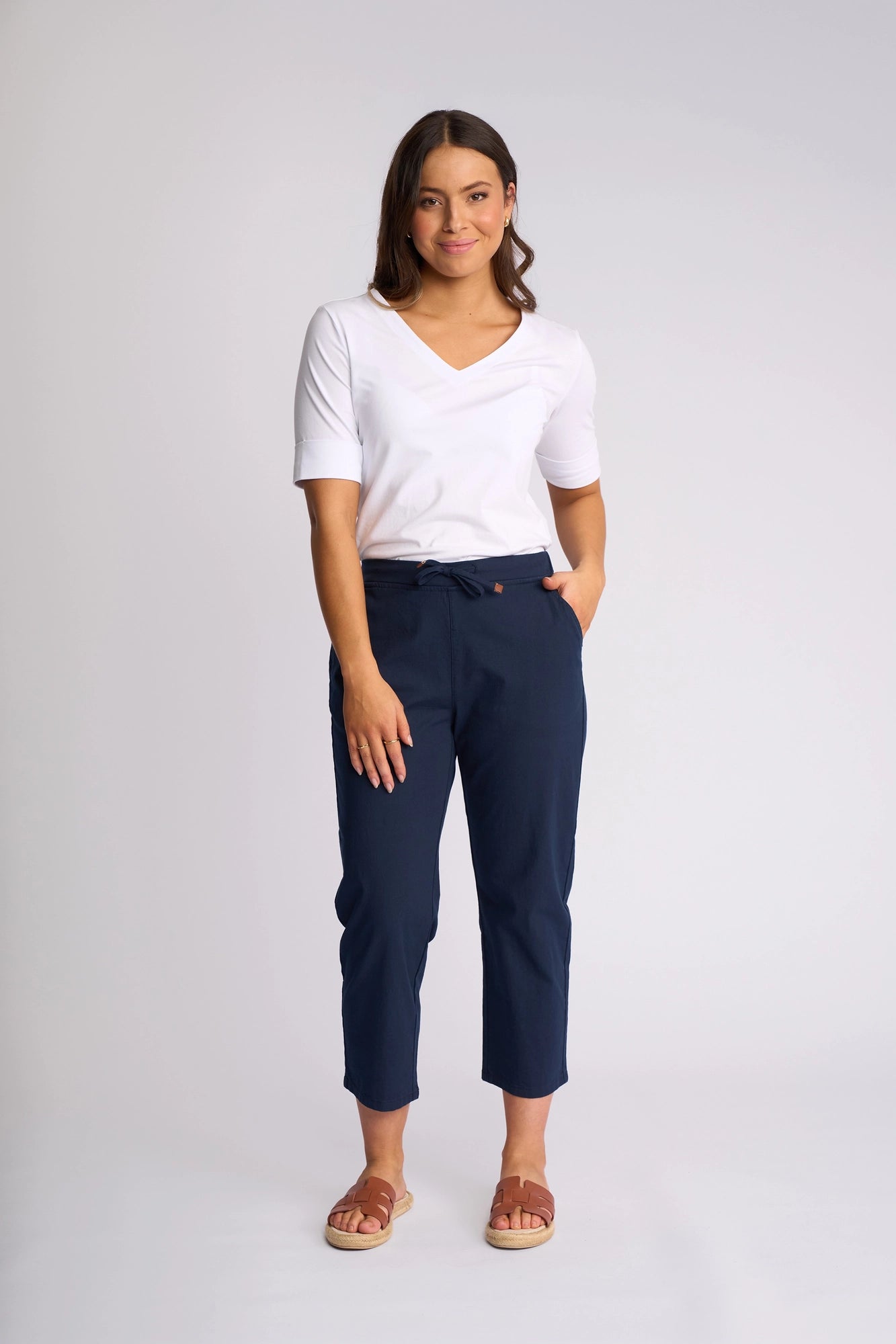 A woman stands against a plain background, wearing a white V-neck top, Vassalli Henley 7/8 Pant in Ink stretch cotton, and beige sandals. She has one hand in her pocket and a slight smile on her face.