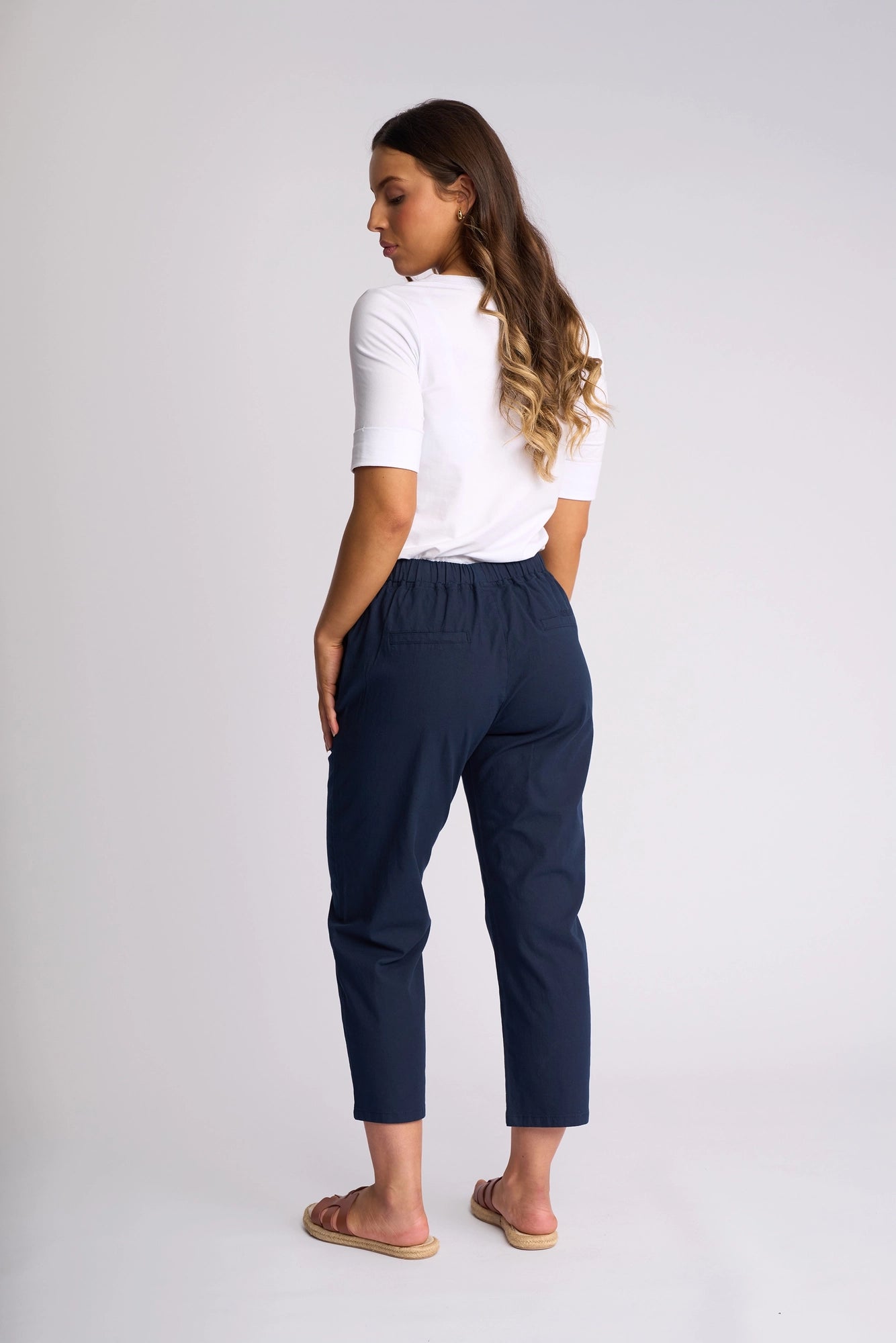 A woman stands facing away against a plain light background, wearing a white short-sleeve top paired with the Vassalli Henley 7/8 Pant - Ink. The stretch cotton pants feature a flattering high waist and modern 7/8 length in navy blue. Her long wavy hair and sandals complete this effortless look.