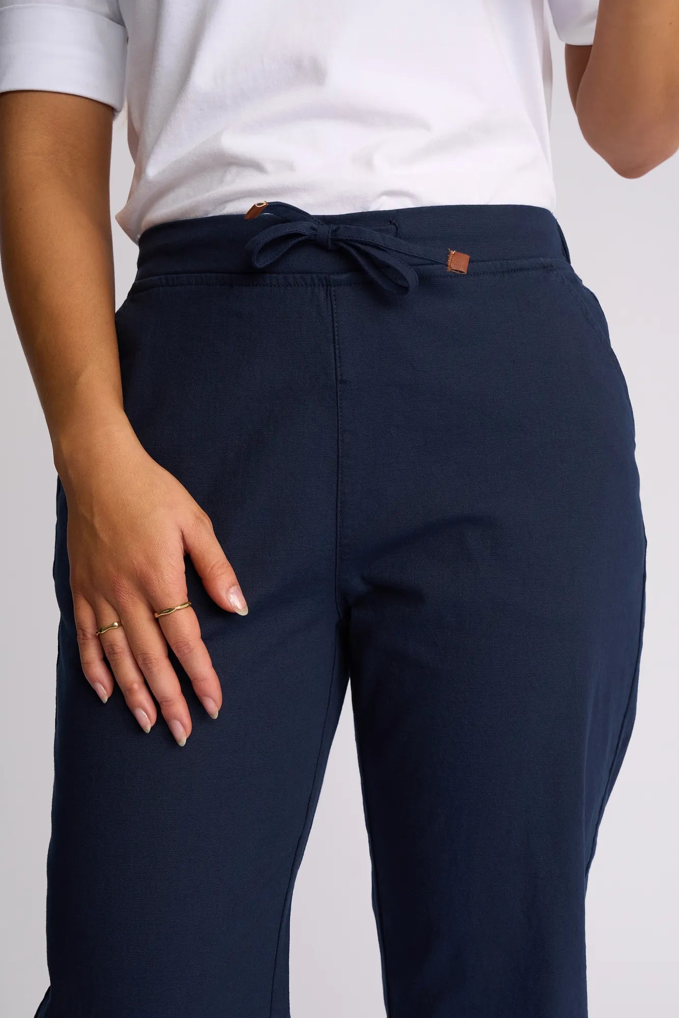 A person wearing a white shirt and Vassalli’s Henley 7/8 Pant in Ink stands with one hand resting on their hip, displaying gold rings on their fingers. Crafted from stretch cotton, the pant offers all-day comfort and ease of movement.