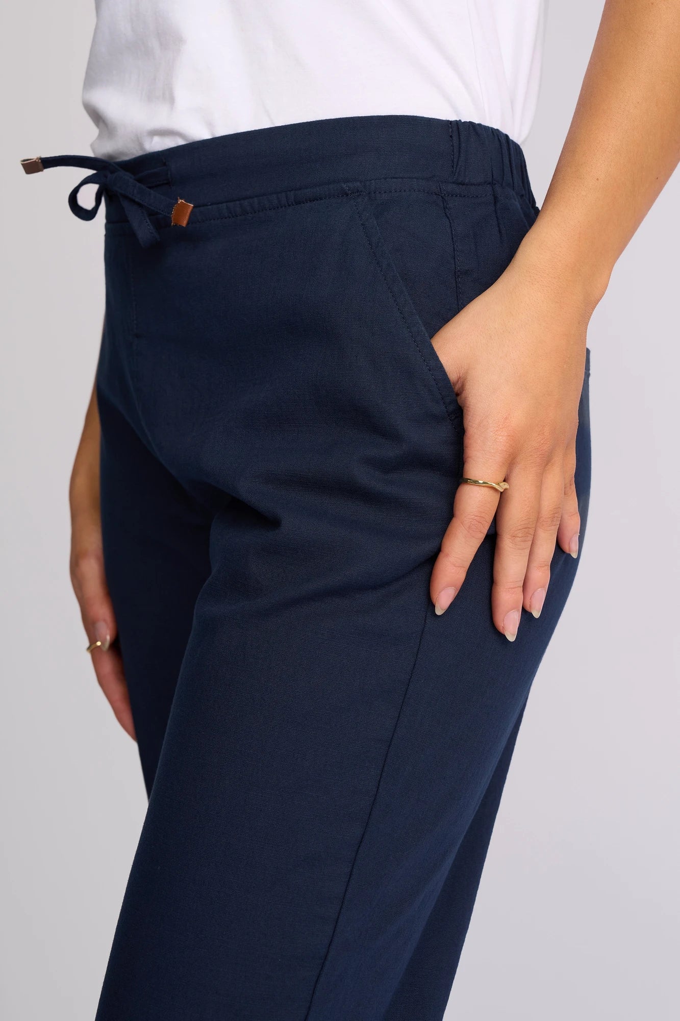 A person wearing the Vassalli Henley 7/8 Pant in Ink, made from stretch cotton, and a white shirt stands with one hand in their pocket, displaying a gold ring on their finger against a plain background.
