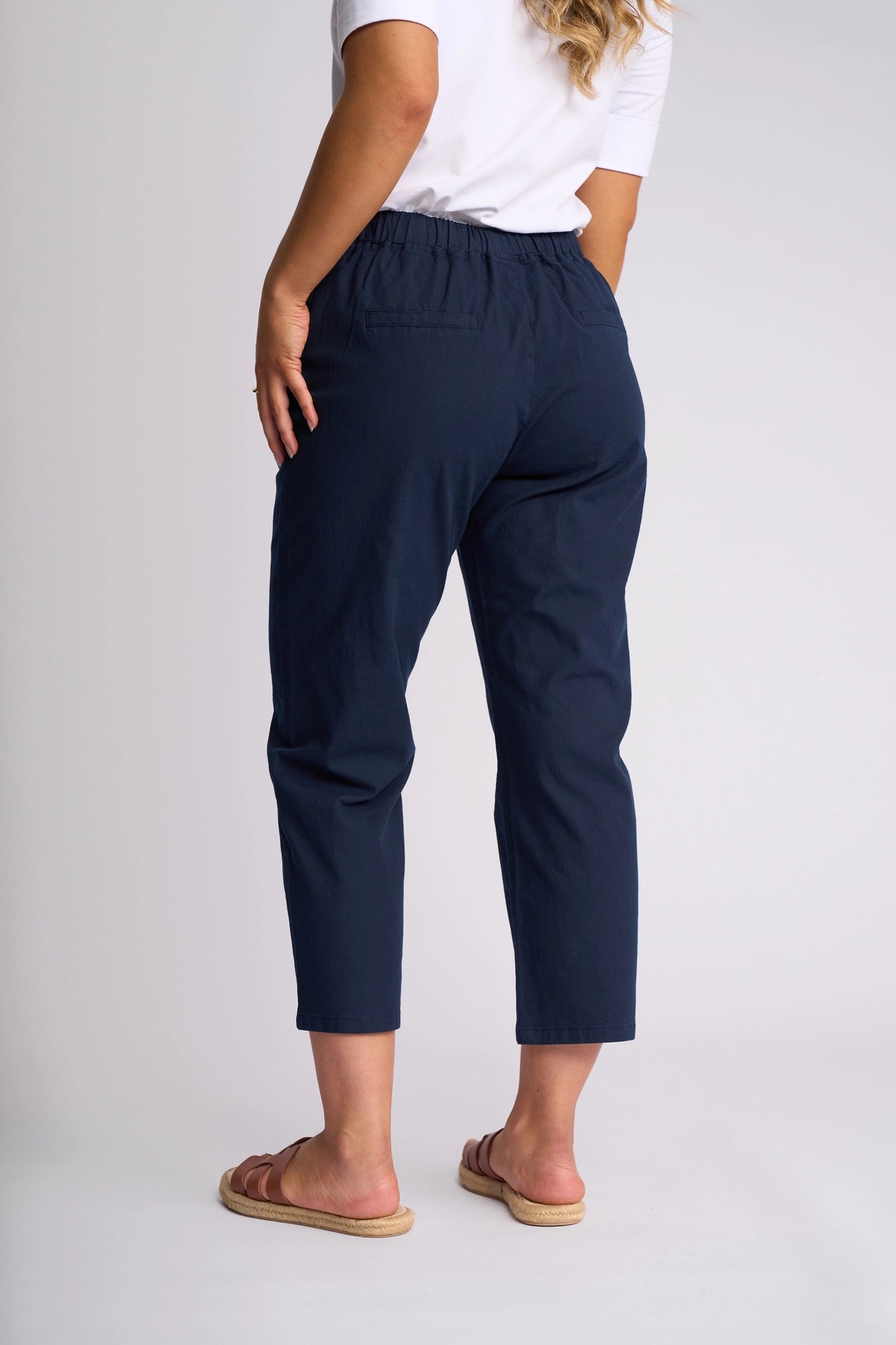 A person stands against a plain background, wearing a white shirt, Vassalli Henley 7/8 Pant in Ink crafted from stretch cotton, and beige sandals. The image highlights the back view of the pant.