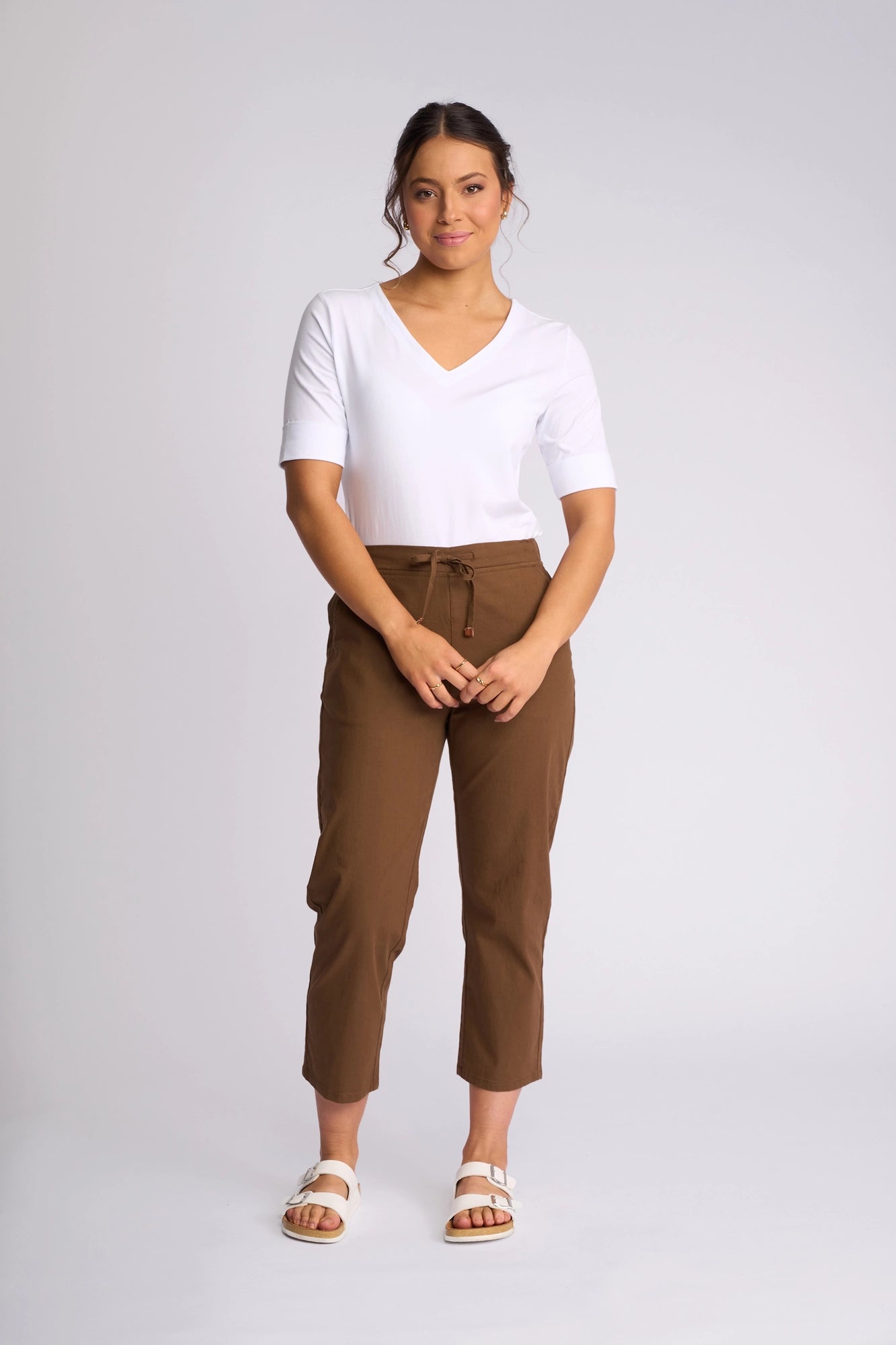 A woman stands against a plain background, wearing a white V-neck short sleeve top, the Vassalli Henley 7/8 Pant in Pecan crafted from stretch cotton, and white slide sandals. Her hair is tied back, and she smiles softly with her hands near her waist.