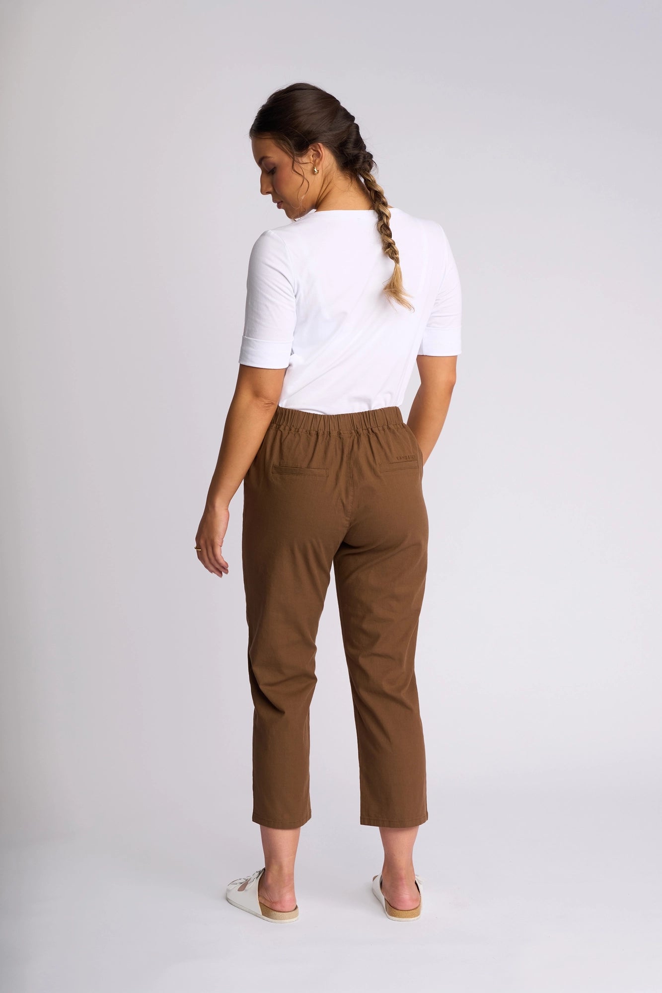 A woman stands with her back to the camera, wearing a white top and Vassalli Henley 7/8 Pant in Pecan, crafted from stretch cotton. She pairs the slim-leg pants with white slip-on shoes. Her hair is braided, and she poses in a studio with a neutral background.