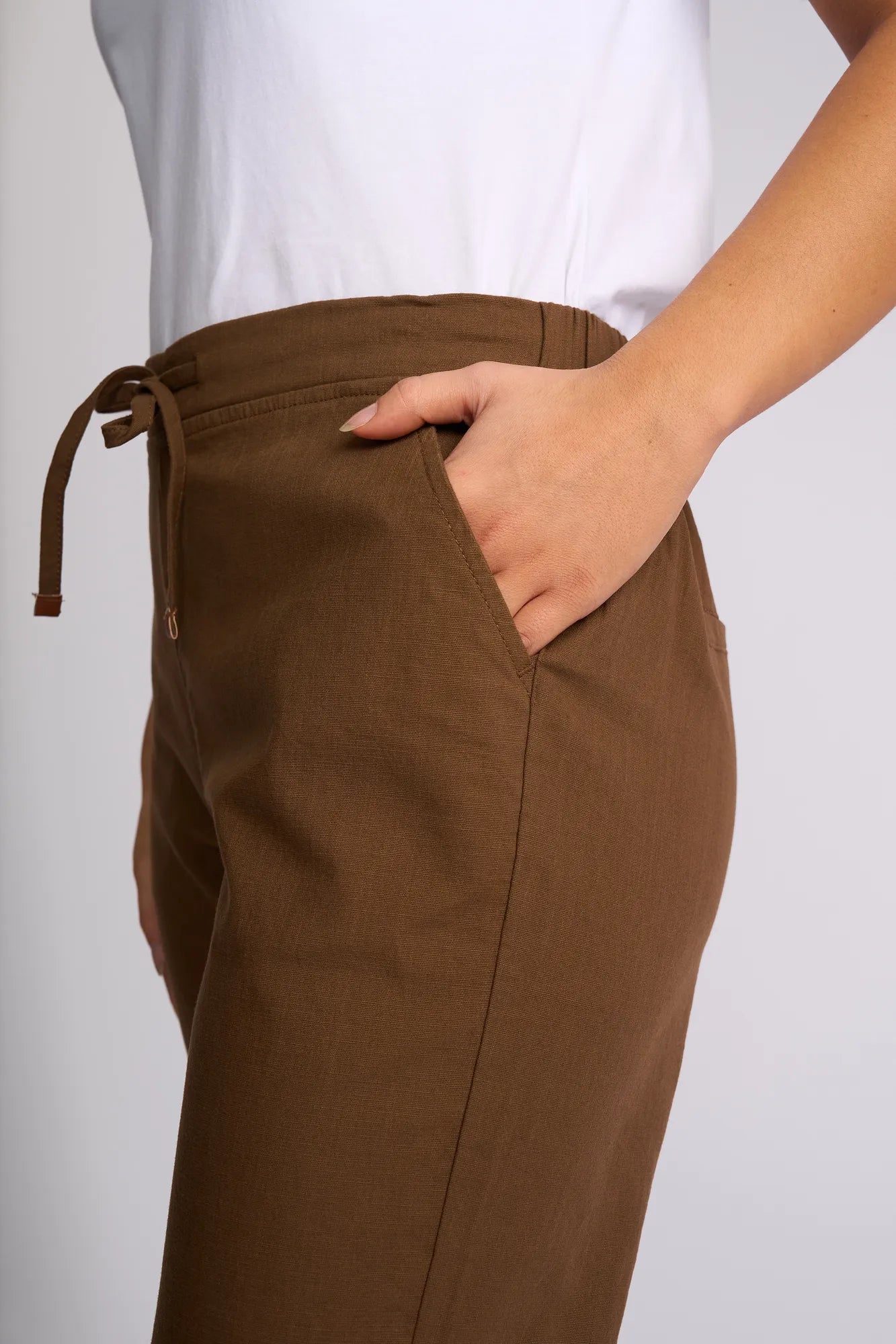 A person wearing Vassalli’s Henley 7/8 Pant in Pecan and a white shirt stands with one hand in their pocket against a neutral background.