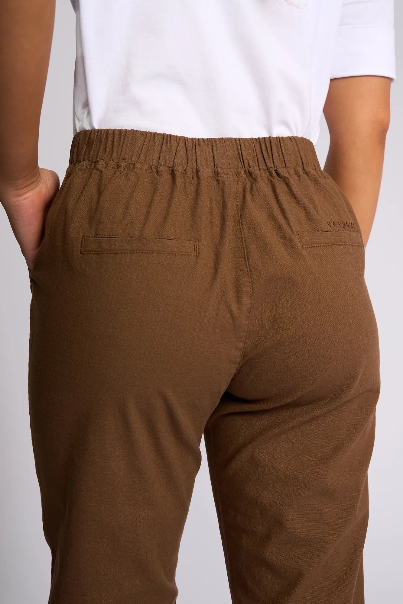 A person is wearing the Vassalli Henley 7/8 Pant in Pecan, featuring an elastic waistband and two back pockets, styled with a white shirt. The photo, taken from the back, highlights the relaxed fit and stretch cotton fabric.
