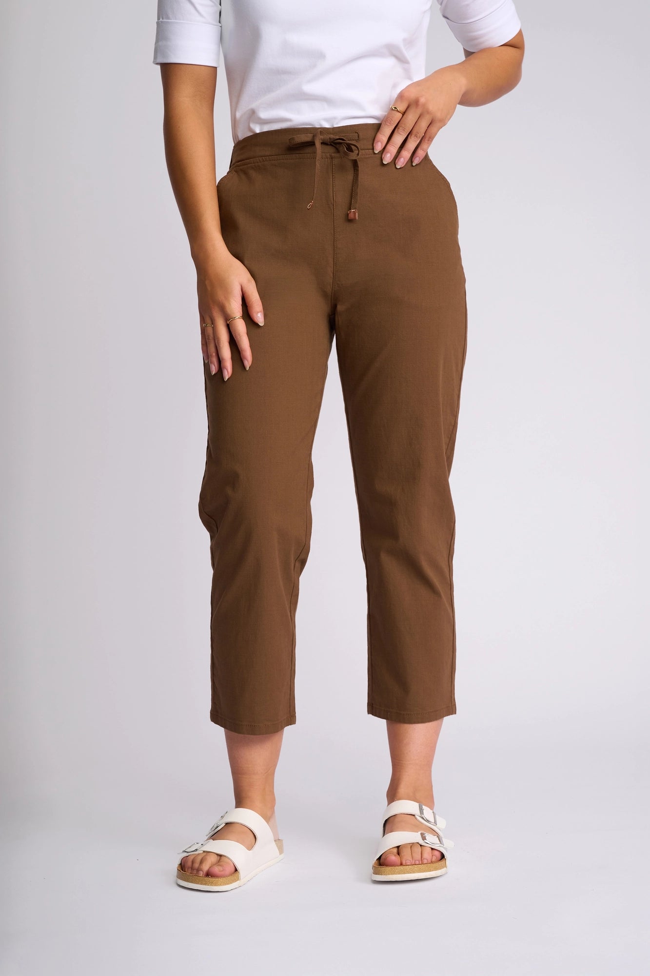 A person wearing a white shirt, Vassalli Henley 7/8 Pant in Pecan—an ankle-length slim-leg style crafted from stretch cotton—and white sandals stands against a plain light background. Only the lower half of the body is visible.