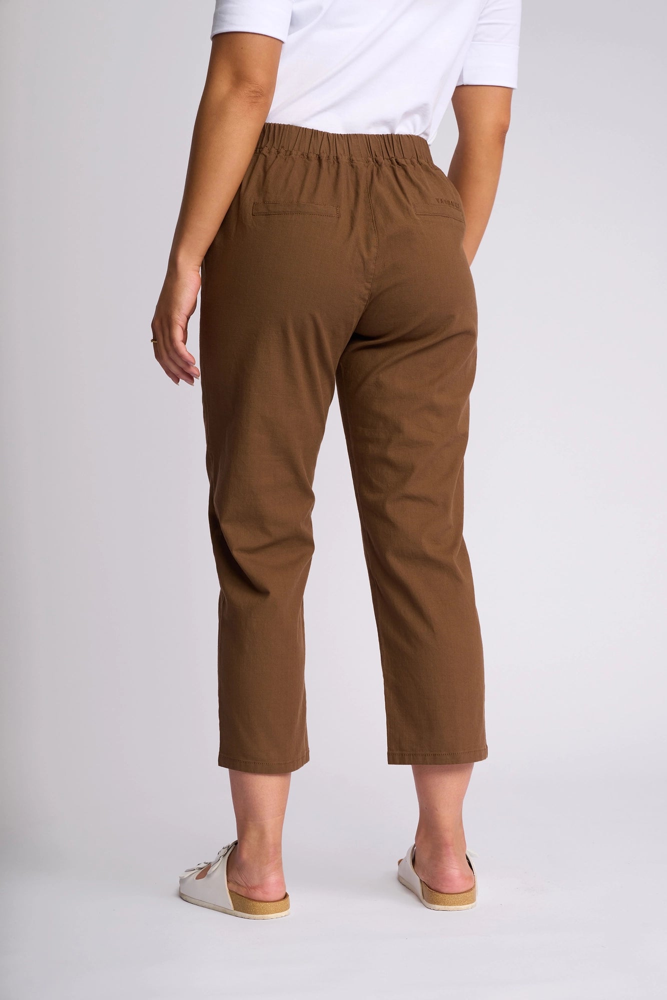 A person is standing with their back to the camera against a plain light background, dressed in Vassalli’s Henley 7/8 Pant in Pecan paired with a white shirt and white slip-on shoes.